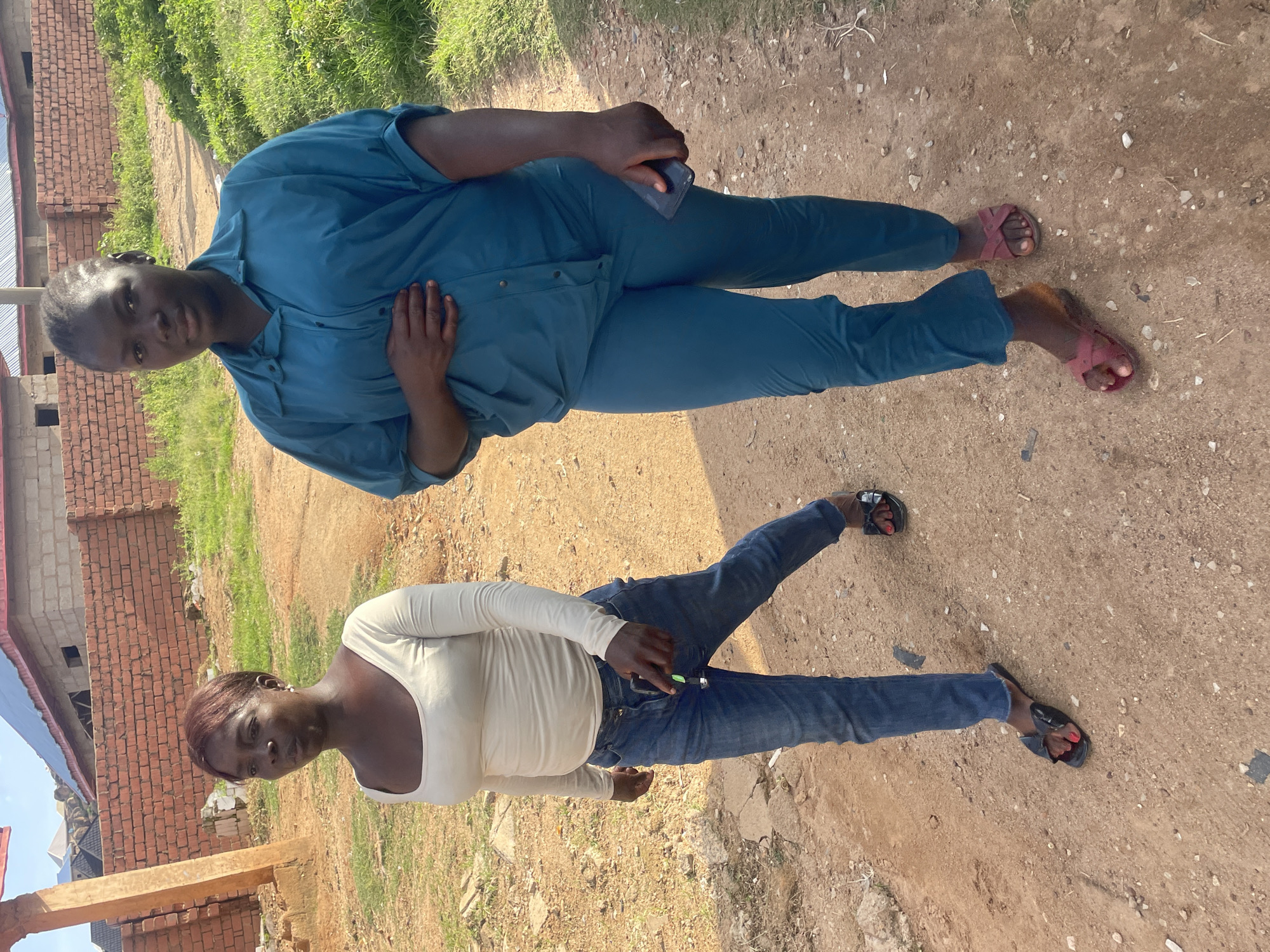 Two women are walking on a dirt path in Barakin Lerawi, Nigeria. The woman in the foreground on the right is wearing a teal-colored shirt and pants, and holding a smartphone in her right hand. The woman on the left is wearing a long-sleeved cream-colored shirt and blue jeans, and appears to be holding a small object, possibly keys, in her left hand. The path is flanked by dry grass and some construction materials, including a brick wall and a wooden post, suggesting an area undergoing development. The sky is visible in the background with some clouds.