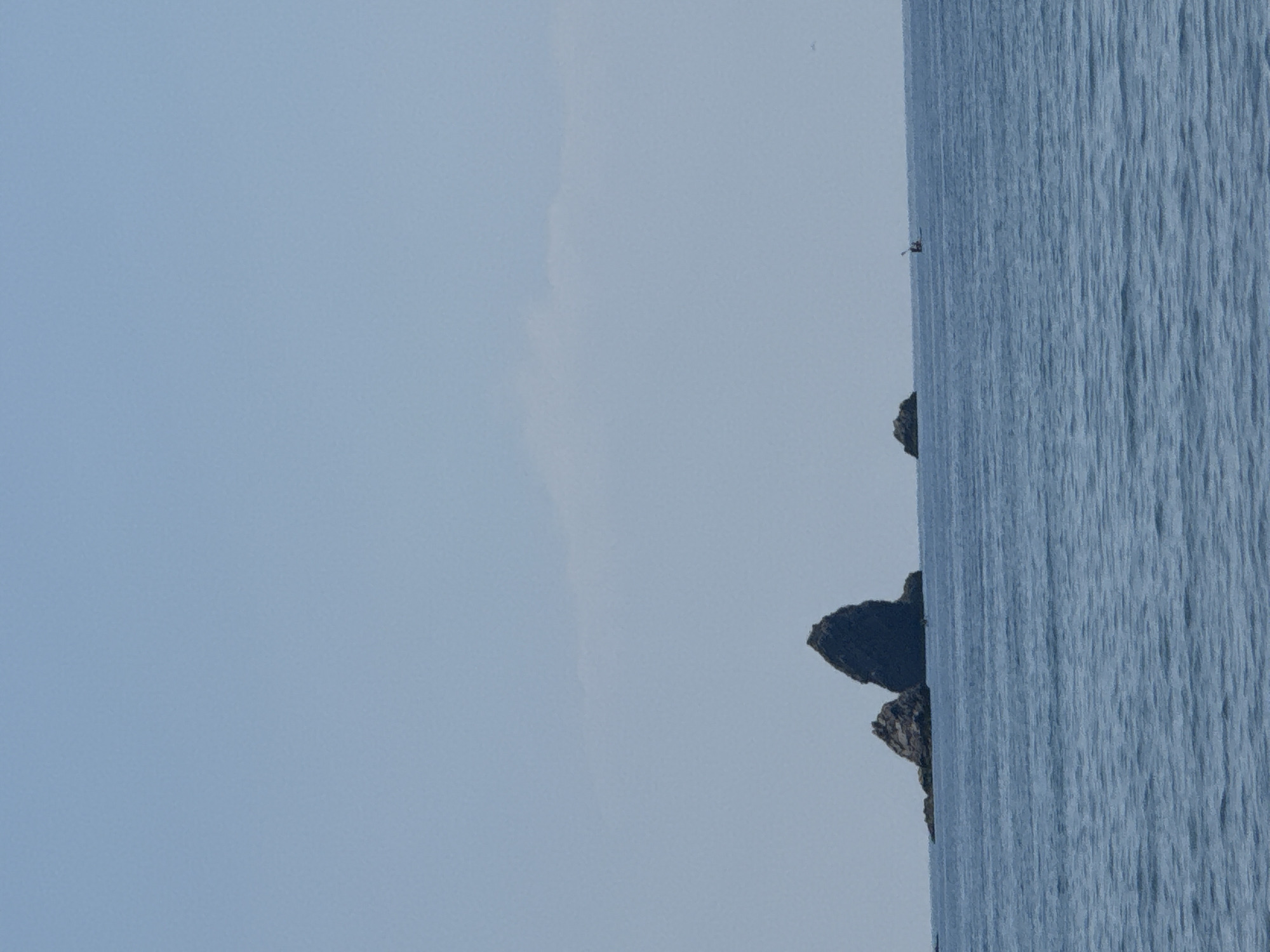 A wide shot shows the calm, blue waters of the Adriatic Sea under a clear, pale blue sky. In the middle ground, three dark rock formations rise from the water. The largest rock is a tall, pointed spire on the right, with a smaller, more rounded rock to its left. To the far right, a small boat with two occupants is visible on the water. In the far distance, a faint cloud formation is visible against the sky. The scene is likely located off the coast of Buljarica, Montenegro, a popular tourist destination known for its natural beauty and coastal attractions.