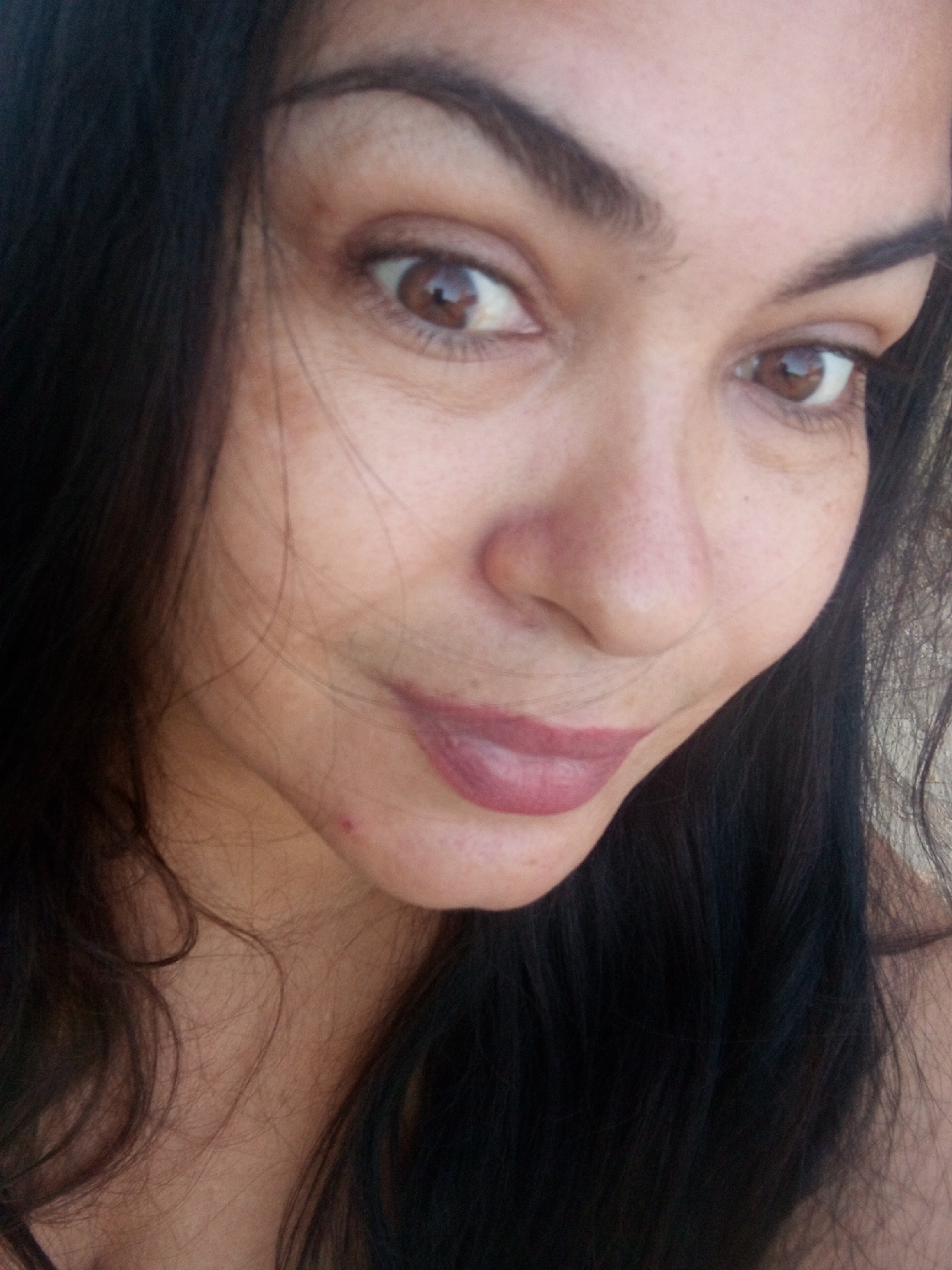 A close-up selfie of a woman with dark hair and brown eyes. Her lips are colored with a mauve-toned lipstick. The background is indistinct, revealing only hints of her hair and shoulder. There are no identifiable landmarks in the image that could definitively place the location in Juan Griego, Venezuela.