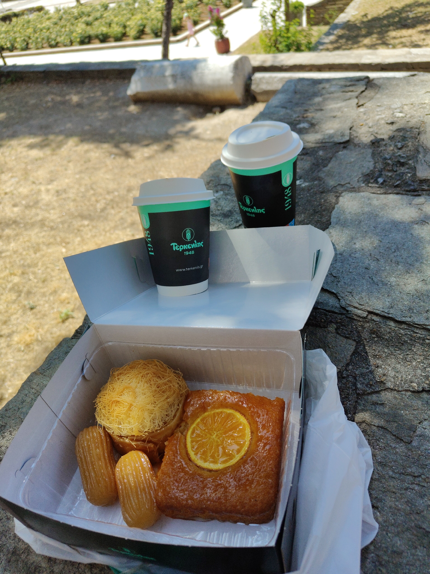 A take-away box of pastries and two coffee cups are situated on a stone surface outdoors. The box contains a round pastry with shredded filo dough, two elongated, ribbed pastries, and a rectangular pastry topped with a slice of candied orange. Two black coffee cups with white lids and green accents, featuring Greek lettering and the year "1948," are placed behind the pastry box. The background shows a blurred park-like setting with green foliage, a pathway, and a large stone. The overall scene suggests a casual outdoor break with refreshments in an urban environment, likely in Thessaloniki, Greece, given the Greek inscriptions on the coffee cups.