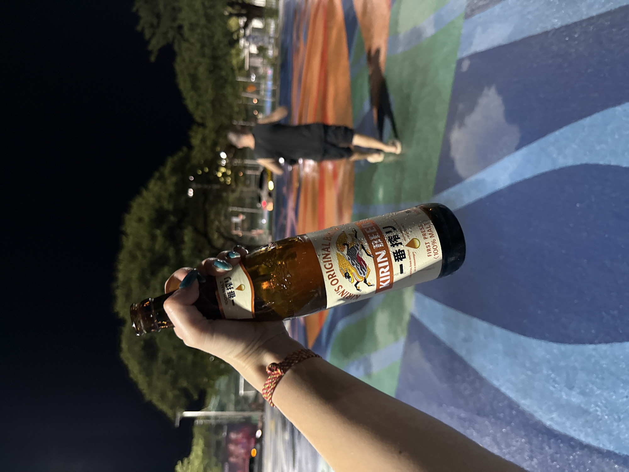 An over-the-shoulder shot shows a person's left hand holding a brown glass bottle of Kirin Beer. The bottle has a yellow and white label with Japanese characters and the words "KIRIN BEER". The person is wearing a red and yellow woven bracelet on their left wrist. In the background, a man in a black tank top and shorts is running across a colorful, multi-colored paved area. Trees and streetlights are visible in the distance. The scene appears to be outdoors at night in an urban or park-like setting. The paved area has a distinctive design with blue, green, and orange sections, suggesting it may be a public recreational space. The overall ambiance is casual and evening-oriented. It is likely the scene is within the city of Ko Samui District, Thailand, given the tropical foliage and outdoor setting.