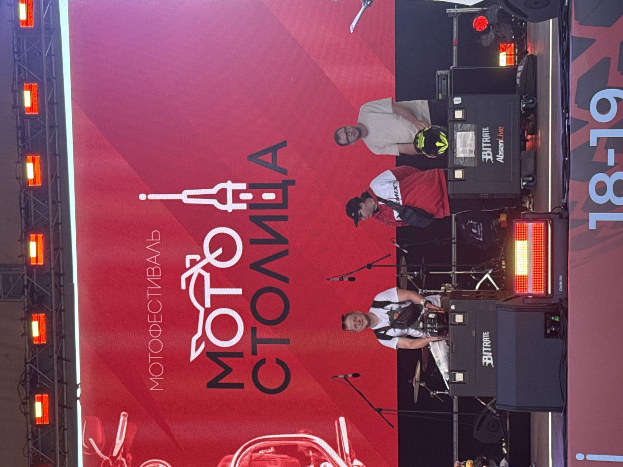 The image captures a segment of an outdoor stage setup, likely for a festival. A large, vibrant red screen dominates the background, displaying the event's title: "МОТОФЕСТИВАЛЬ" (Motofestival) at the top, followed by a stylized logo of a motorcycle and the words "МОТО СТОЛИЦА" (Moto Capital). Three men are visible on stage. The man on the left is wearing a white t-shirt, a black fanny pack, and sunglasses. He is seated behind a drum kit, suggesting he might be a performer. The middle man is wearing a red and white hooded sweatshirt with "EMIX" written on it, a black crossbody bag, and a black baseball cap. The man on the right is wearing a light-colored t-shirt, glasses, and is holding a black and yellow motorcycle helmet. Two large black speaker cabinets with "BITRATE" and "AbsenLive" logos are positioned on the stage. Behind the men, various stage equipment, including microphones, cables, and lighting rigs, are present. The overall setting indicates a public event, potentially a motorcycle festival, possibly taking place in Saint Petersburg, Russia, given the Cyrillic text on the screen. The bottom of the image shows a red banner with white text "18-19", indicating dates for the event.