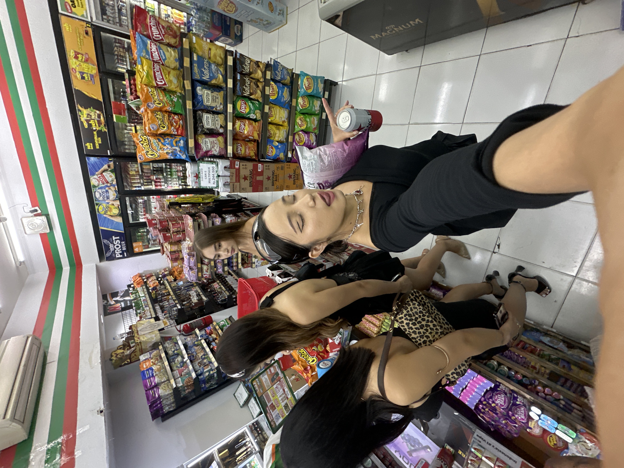 **Setting:** Interior of a convenience store.
**Location:** North Kuta, Indonesia is suggested by the Indonesian text, products, and prices.
**People:** Three young women are present. One woman is holding a can of drink in a selfie-style photograph. The second woman carries a leopard-print bag.
**Objects:** Shelves stocked with a wide variety of snacks, including chips (Cheetos, Lay's, Crunchips, natural chips), beverages, and various convenience store items. An air conditioner is visible near the left side of the frame. A red shopping basket is visible.
**Actions:** The women are shopping inside the convenience store. One woman is taking a selfie with a can in hand.
**Environment:** The store is brightly lit. The products and signage indicate a retail setting. The walls are decorated with red, green, and white stripes. The layout suggests a typical convenience store with shelves and aisles.
**Additional details:** Brands such as "Bintang" beer are visible. Text is on the shelves.