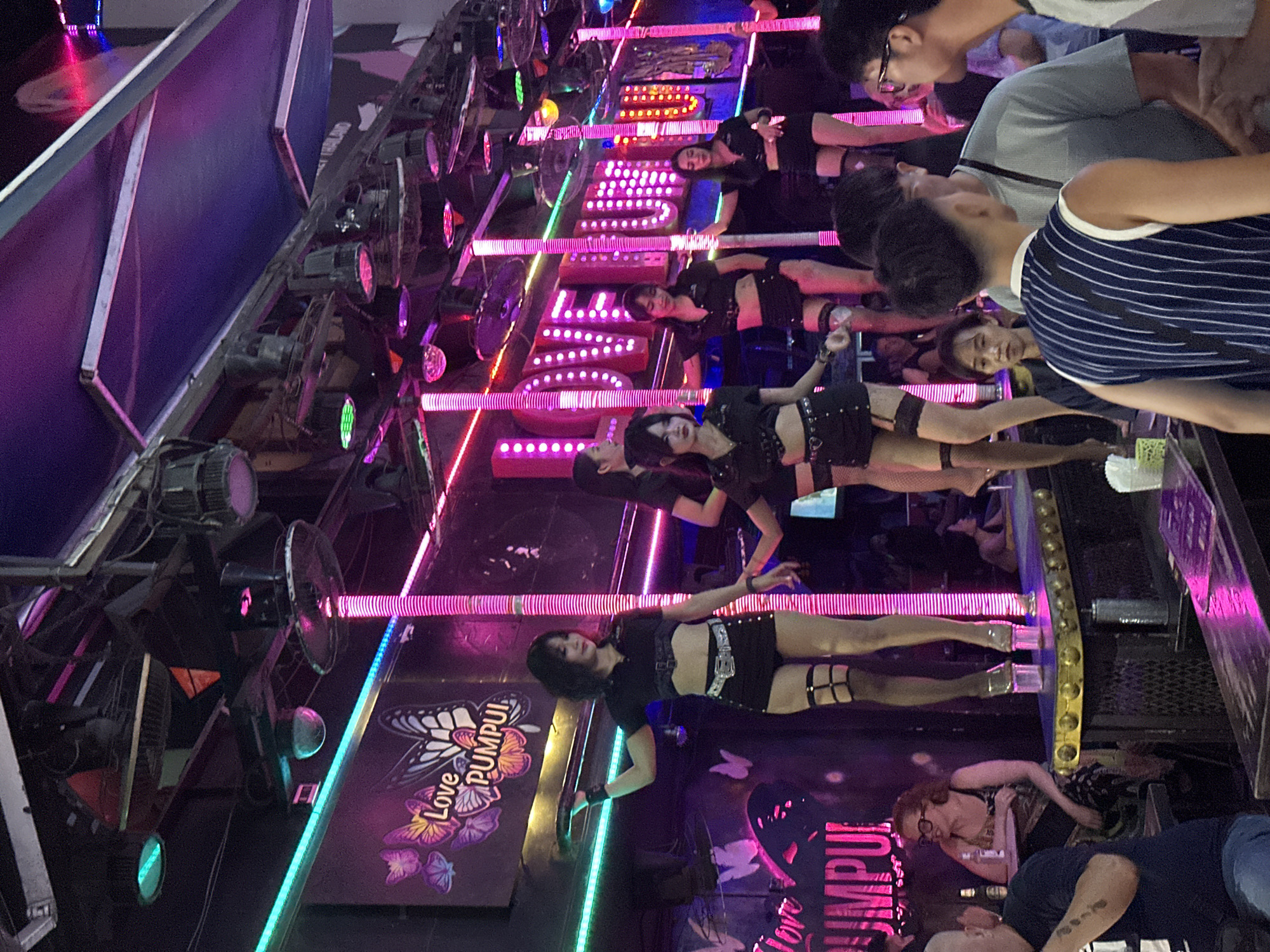 The image depicts an open-fronted entertainment venue at night in Patong, Thailand. The scene is dominated by vibrant neon lighting in pink, magenta, blue, and cyan. Overhead, the ceiling structure supports multiple spotlights, fans, and what appear to be audio speakers. Large illuminated signs prominently display "Love PUMPUI" with butterfly graphics.
On an elevated stage or platform, approximately six women are actively performing. They are uniformly dressed in black short-sleeved crop tops, black shorts, black thigh-high stockings with garter belt straps, and clear platform heels. Several women are posing or dancing, some leaning against or holding onto vertical illuminated poles that extend from the stage.
In the foreground, a group of male patrons is partially visible. One individual wears a blue and white striped sleeveless shirt and faces away from the camera. Another man in a light grey t-shirt stands nearby. To the left, a male patron with a shaved head is seated at a table. Behind him, a woman with red hair and glasses, wearing a patterned dress, is seated at a table and smiling. Other patrons are discernible in the background, seated or standing, observing the performances. A counter with a white tissue dispenser and printed material is visible in the immediate foreground.