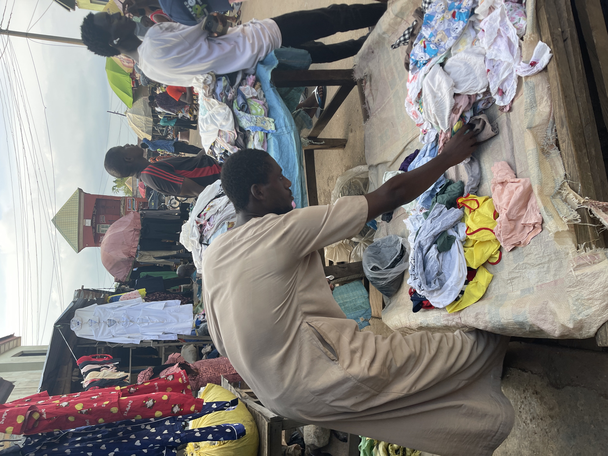 The image shows a market scene in Akwanga, Nigeria. A man in a tan garment is seated, examining clothing on a table. A second person is seated behind him, with more clothing in piles on the table. In the background, more people are visible amidst market umbrellas and stalls. Other clothes and clothing items are hanging from racks. The environment suggests a bustling open-air market.