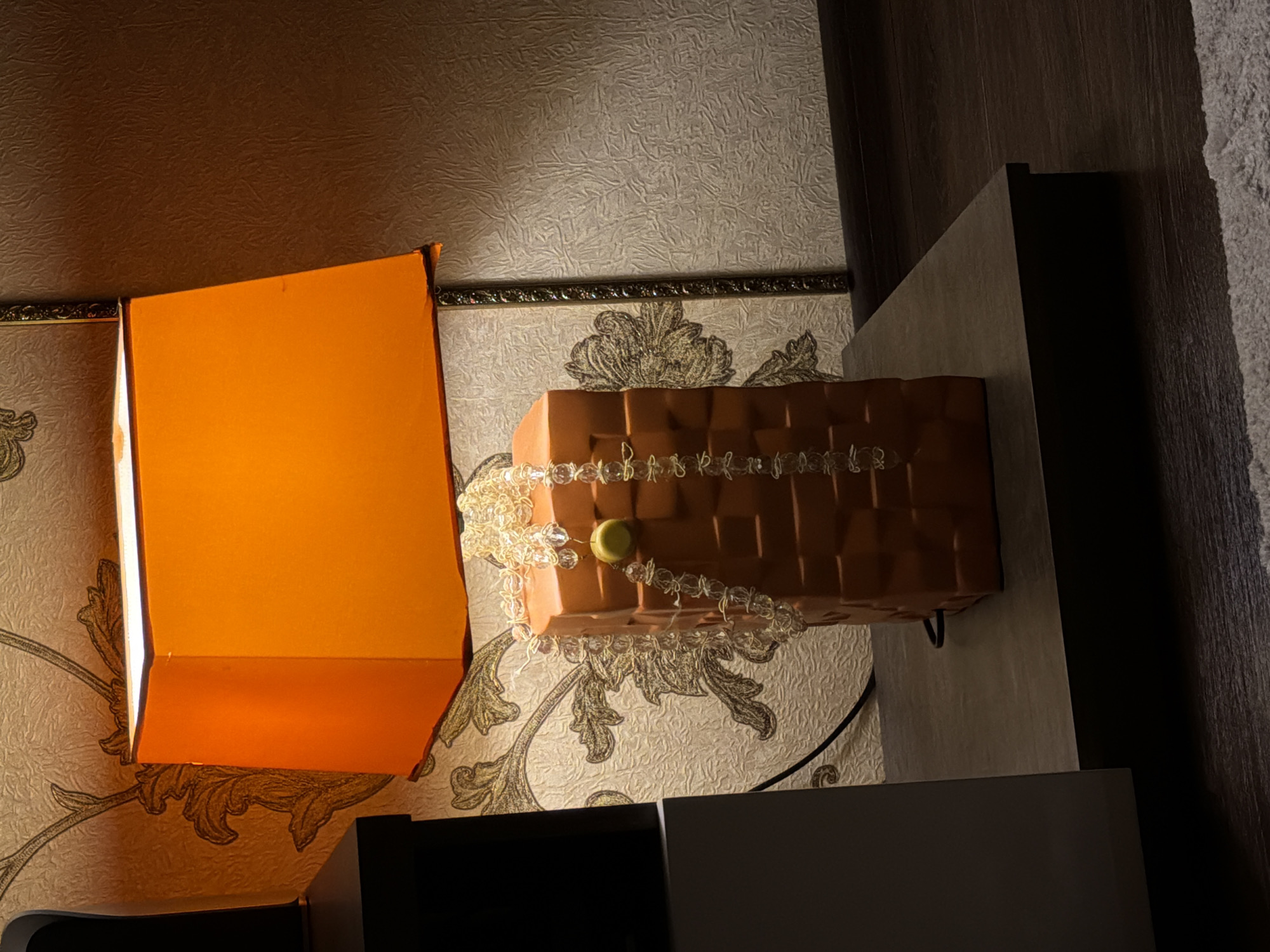 A high-angle shot depicts a room in Penza, Russia, featuring a patterned wallpaper, an orange lampshade, a decorative brick-shaped object adorned with a beaded garland, and a dark-toned cabinet. The lampshade emits a warm glow, illuminating a portion of the wall. The cabinet's surfaces reflect ambient light, creating tonal variations. A black wire is visible, likely associated with the lighting fixture. The overall impression is that of a furnished interior space with a mix of functional and decorative elements.