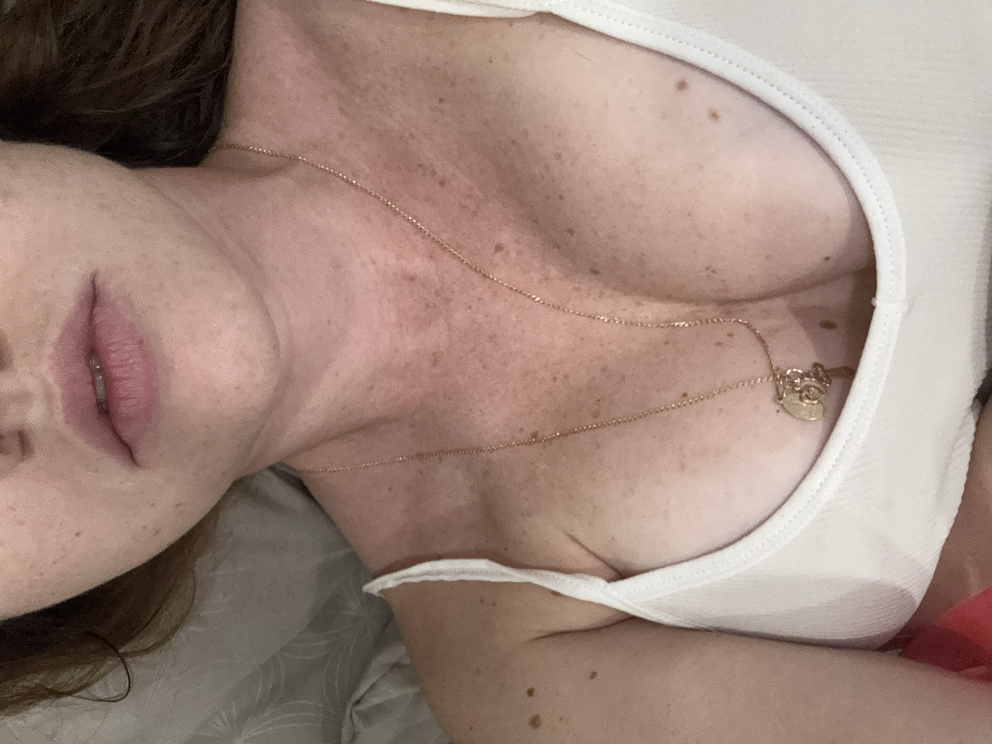 A close-up, indoor, overhead shot focuses on a person's chest and neck area, revealing fair skin with freckles. The person is wearing a white ribbed tank top, with a delicate gold necklace featuring charms resting on their décolletage. The background consists of a rumpled grey textured surface, possibly bedding. The image is a still from an unspecified recording, and no specific location in Rawai, Thailand, can be identified from the visual information provided.