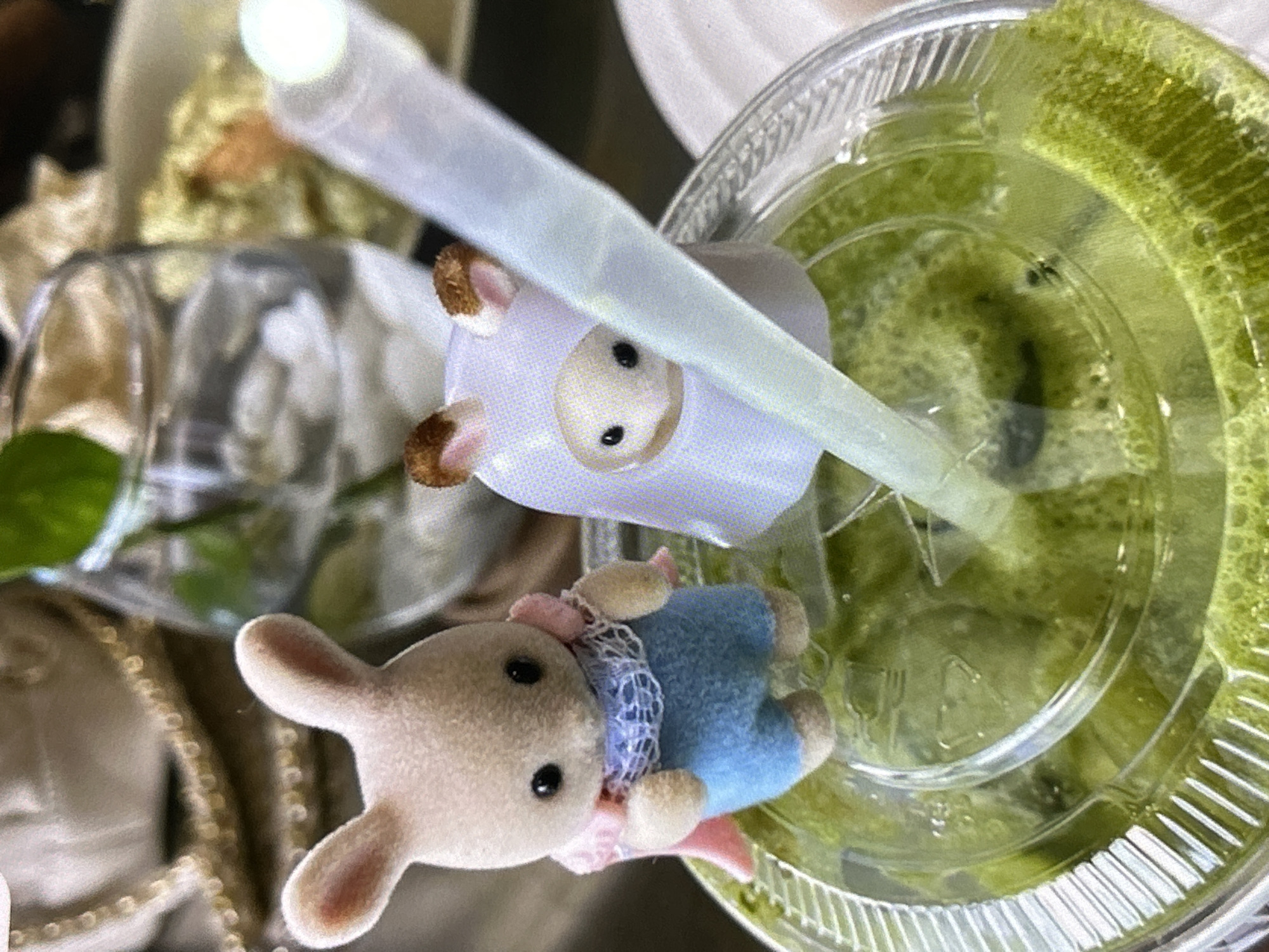 A close-up shot shows two small plush toys in front of a plastic cup of green liquid, likely a beverage. The toy on the left is a beige bunny wearing blue overalls and a pink cape with white trim. The toy on the right is a white ghost-like figure with brown ears and a rounded face with black eyes and a small mouth. A clear plastic straw is positioned between the two toys, extending into the green liquid. The background is blurred, with hints of a glass of water with ice cubes, and a table setting that suggests a cafe or restaurant. There are no visible people or discernible actions. The lighting is natural, with a bright light source in the upper right corner. The image appears to have been taken indoors. The location in Depok, Indonesia is not specified.