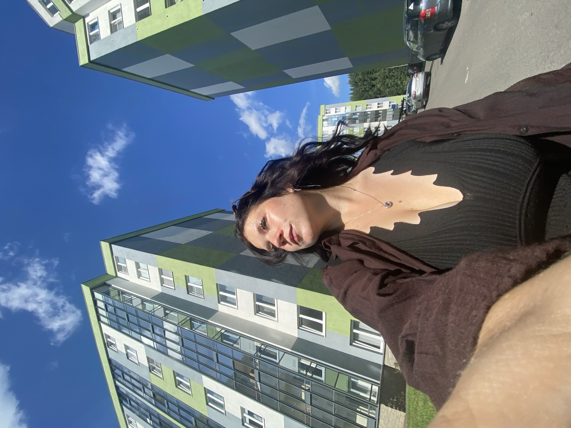 In Yanino-1, Russia, under a bright blue sky with scattered white clouds, a young woman stands in front of two modern, tall apartment buildings. The buildings feature a distinctive checkered facade of light gray and lime green. The woman, with long, dark, wavy hair, is looking upwards and to her left, her face illuminated by sunlight. She is wearing a dark, ribbed top with a deep V-neck and a brown, open button-up shirt over it. A delicate silver necklace with a circular pendant adorns her neck. In the background, a paved road runs between the buildings, with several cars parked or driving. Green grass covers the ground in front of the buildings. The perspective of the photo is low-angle, emphasizing the height of the apartment buildings and the expanse of the sky.