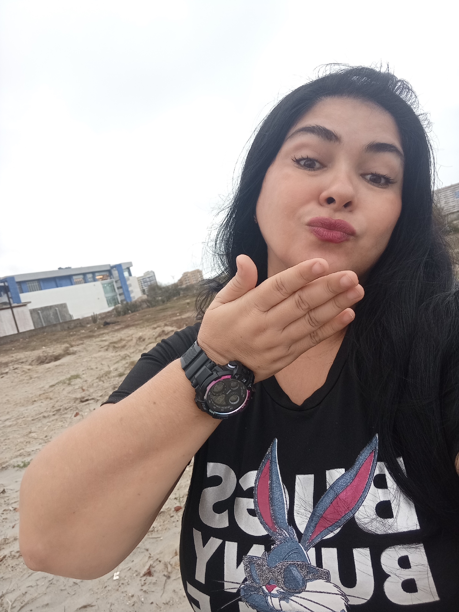 A woman with long, dark hair and dark eyes, wearing a black t-shirt with a Bugs Bunny graphic, poses for a selfie outdoors. She is puckering her lips and extending her left hand towards the camera, as if blowing a kiss. A digital watch with a black band is visible on her left wrist. In the background, there is a sandy, uneven terrain with a modern building featuring blue and white elements, and a grey wall. The sky is overcast and bright.