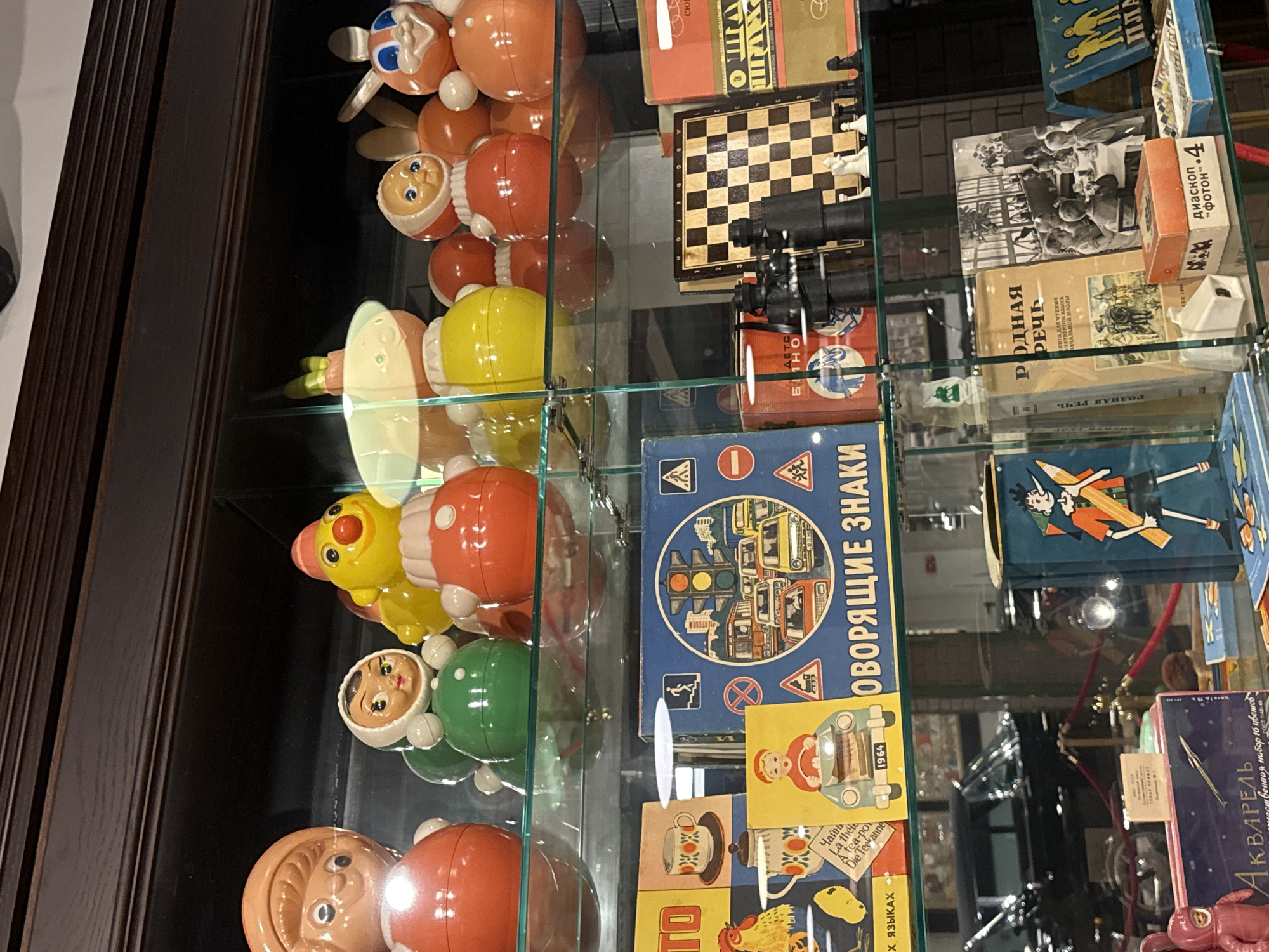 This image shows a display case at the Arkhangelskoye Estate Museum in Arkhangelskoye, Russia, featuring a collection of vintage toys and books.
On the top shelf, several "Nevalyashka" (roly-poly) toys are visible, including a large orange one with a girl's face on the left, a smaller one with a green body and a girl's face, and a yellow chick-like toy with a red nose. Other similar toys with red and yellow bodies are also present.
The middle shelf contains various items. On the left, there are brightly colored boxes with illustrations, one depicting a rooster and another with a teacup and text in multiple languages. A blue box with traffic-related illustrations and the Cyrillic text "ОПОРЯЩИЕ ЗНАКИ" (warning signs) is prominently displayed. Next to it is a chessboard with chess pieces set up. To the right of the chessboard, a red box with illustrations and text, likely a game or puzzle, is visible.
The bottom shelf holds a collection of books and other objects. A dark blue box with an illustration of a boy with a long nose, possibly Pinocchio, is on the right. To its left is a book with the Cyrillic title "РОДНАЯ РЕЧЬ" (native speech), accompanied by a black and white photograph of children gathered around a table. Several smaller boxes and what appear to be toy binoculars are also on this shelf. In the background, parts of a car and glass barriers are visible, suggesting a museum setting. The overall environment is an indoor display case with controlled lighting.