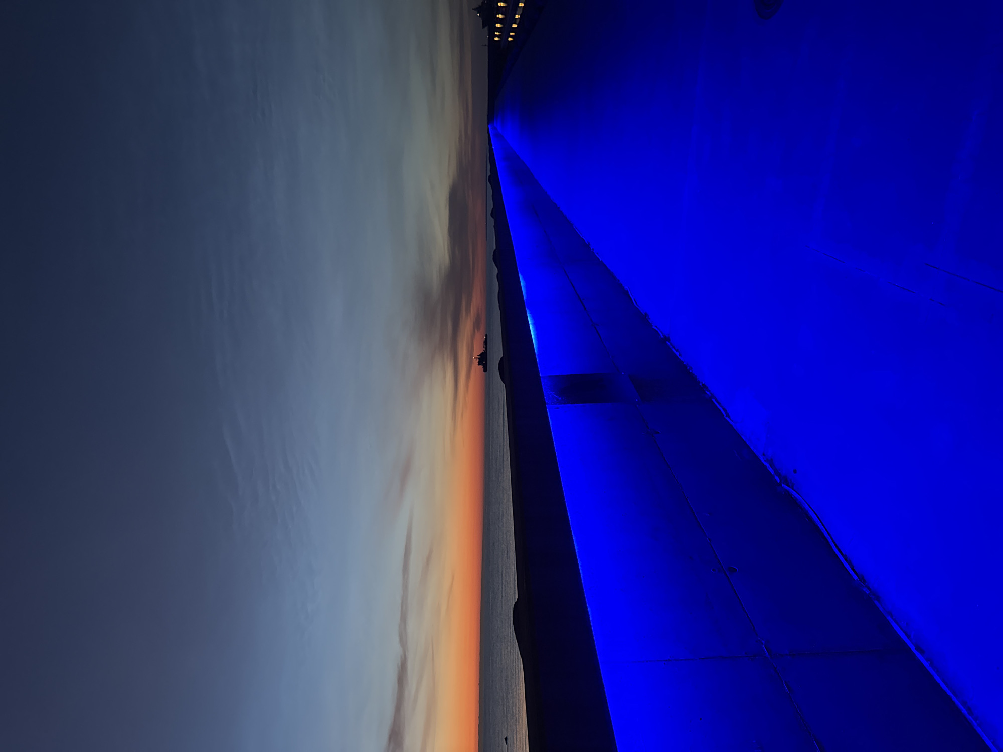 The image captures a sunset or sunrise over the Baltic Sea from a pier in Tallinn, Estonia. The sky transitions from deep blue at the top to vibrant orange and yellow near the horizon. Several ships are visible in the distance on the water. The foreground of the image is dominated by a walkway or pier illuminated by bright blue LED lights, creating a striking contrast with the natural light of the sky. The pier appears to be a modern structure, possibly a promenade or harbor infrastructure. The lighting suggests it is either dusk or dawn, with the artificial lights of the pier becoming prominent as natural light fades.
