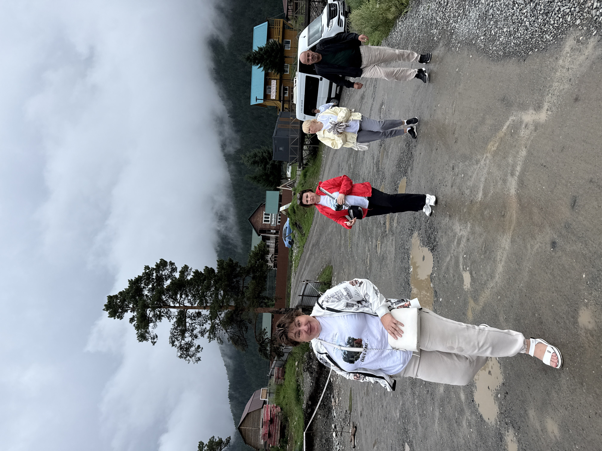 The image depicts a group of four people walking on a muddy, unpaved road in Elekmonar, Russia. The weather is overcast and foggy, with mist obscuring the forested hills in the background. The foreground features a woman in a white jacket and white pants, holding a white purse. Behind her, a woman in a red jacket walks away from the camera, followed by a woman in a yellow jacket and a man in a dark jacket. A white van is parked to the right of the group. Several wooden buildings, including houses and what appears to be a shed or storage area, are visible in the middle ground. The overall scene suggests a rural or remote location, possibly a tourist destination, with signs of recent rain evident in the puddles on the road.
