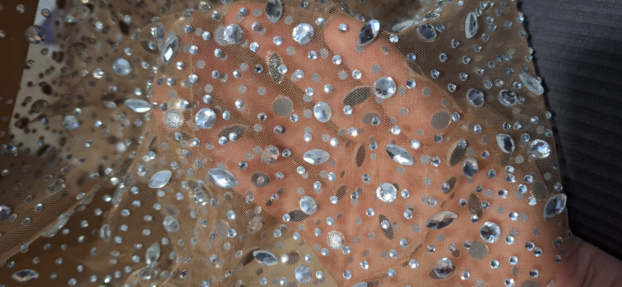 A close-up, high-angle shot features a piece of sheer, light brown or nude-colored mesh fabric adorned with numerous clear, sparkling rhinestones and smaller silver embellishments. A human hand, likely an adult's, is visible underneath the translucent fabric, serving as a skin-toned background.
The rhinestones vary in size and shape; there are many small, round faceted crystals, along with larger, more prominent oval, teardrop, and round shapes. These crystals reflect light, creating bright highlights across the fabric. Scattered among the clear rhinestones are smaller, flat, silver-toned embellishments, some appearing circular and others oval. The fine mesh texture of the fabric is discernible through the pattern of the attached adornments. On the right side of the frame, the embellished fabric meets a darker, possibly grey or black, solid material or surface. The image emphasizes the intricate detailing and shimmer of the textile. No activity other than the fabric being held is depicted. There is no visible text.