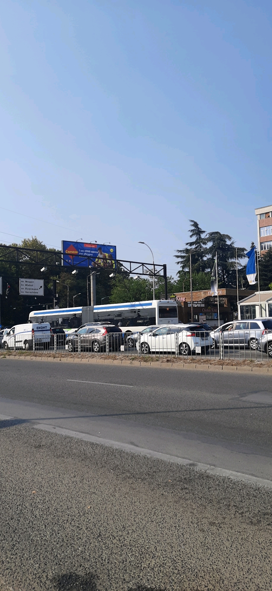 This is a photograph of a street scene in Varna, Bulgaria, on a clear day. The image captures a traffic jam with several cars and a bus lined up on the road. In the background, a large billboard is visible, featuring an advertisement for a lottery with the text "Спечели 1 от 4000 монки". There are also traffic signs indicating directions to "кв. Младост", "кв. Възраждане", and "кв. Vazrazhdane". A building with "Chicken King" signage is also present, along with flags. The foreground shows a sidewalk and the road. The sky is clear and blue.