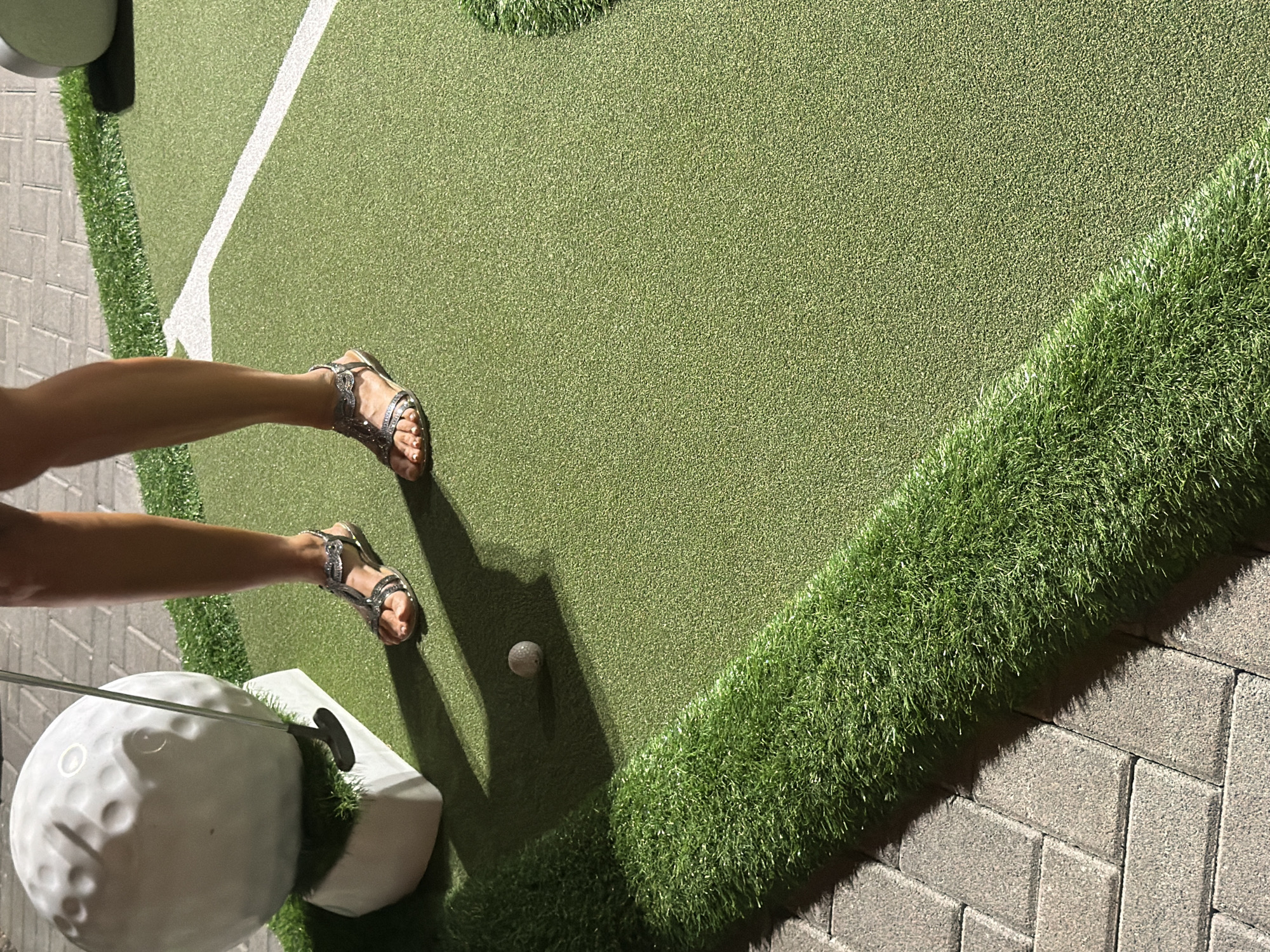 In Gatteo, Italy, a person's legs and feet, adorned with decorative sandals, are visible as they stand on a miniature golf course. The individual is positioned near a large, white golf ball, which rests on a white pedestal. A golf club is being held by the person, poised to strike the ball. The ground is covered in green artificial turf, with a white line marking a boundary. To the left, a portion of a paved walkway with grey rectangular bricks is visible. The scene is lit by artificial lighting, casting shadows.
