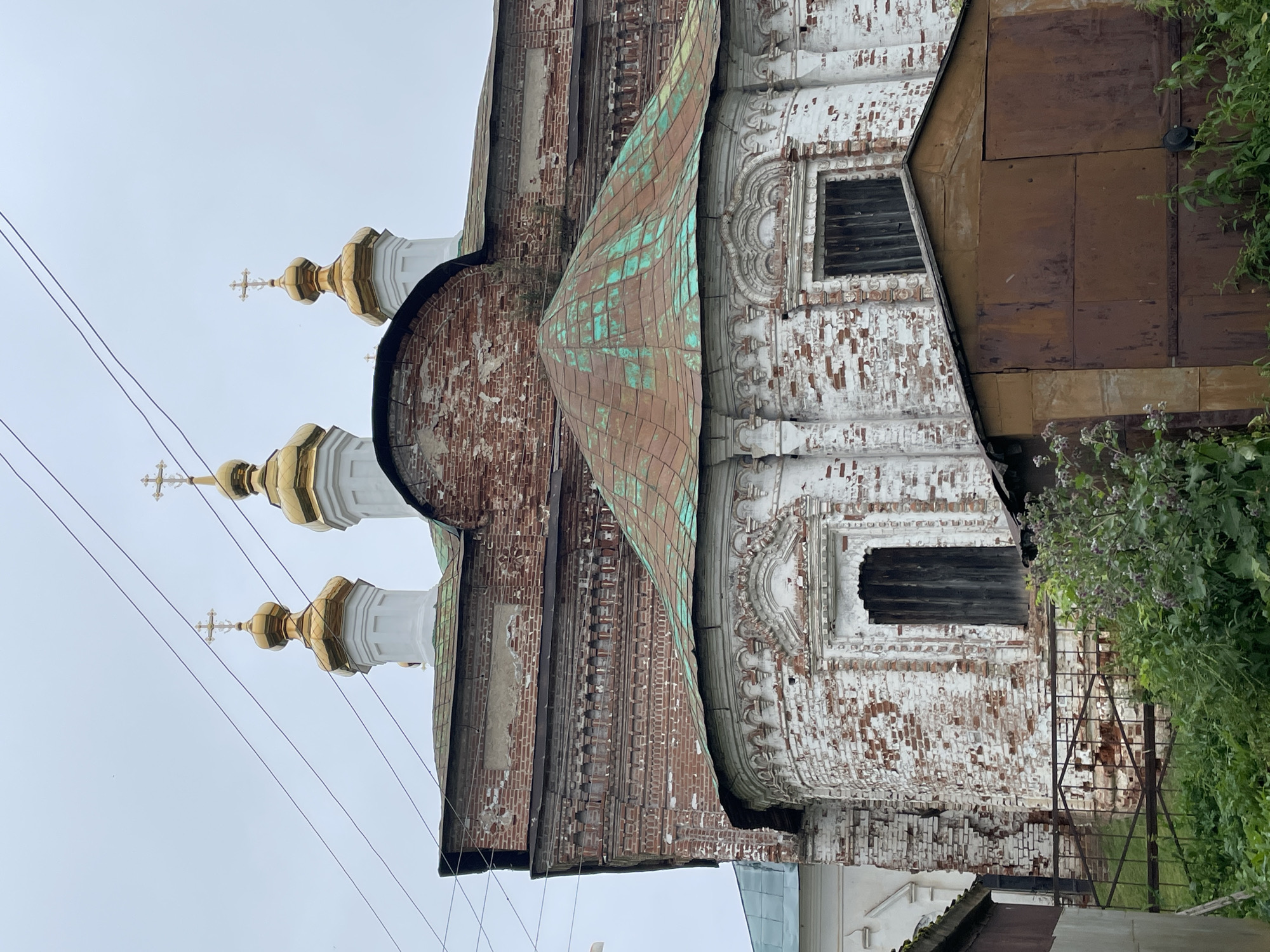 This image depicts the Trinity Church in the village of Kstinino, Russia, under an overcast sky. The church features a red brick exterior with white decorative elements, showing signs of age and weathering. Three golden onion domes with crosses are visible at the top, supported by white columns. The church has a green copper roof that is visibly oxidized. In the foreground, a rusty metal garage or shed stands, partially obscuring a metal fence and lush green vegetation. The overall impression is one of a historic religious building in a rural setting, showing the passage of time.