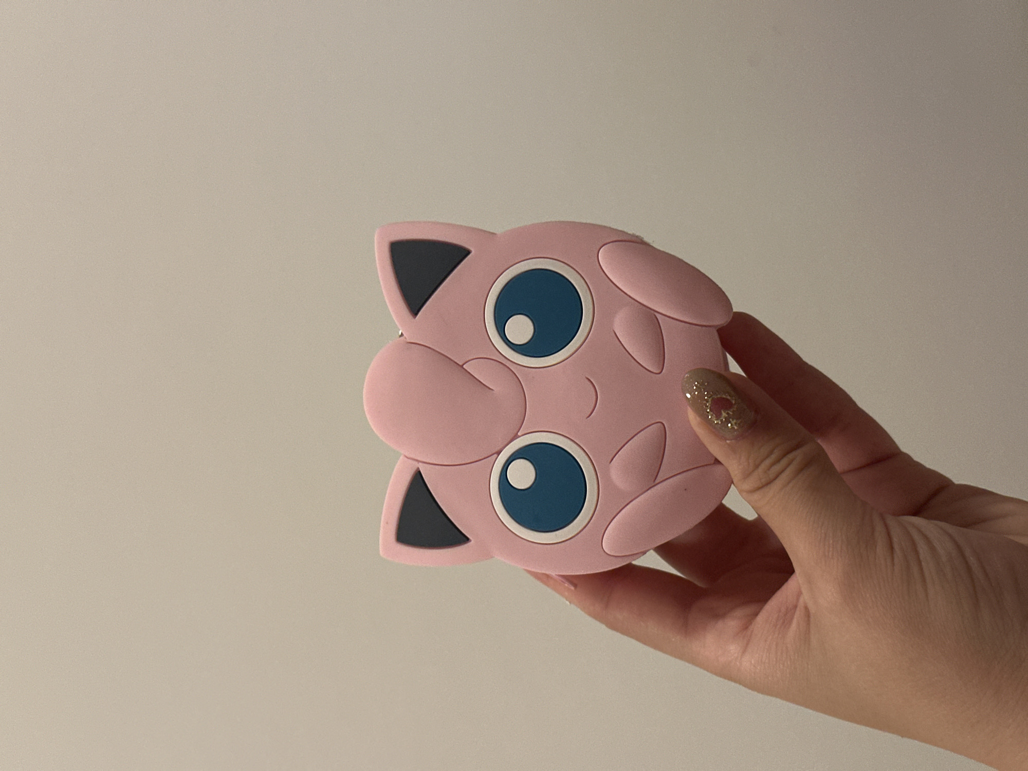 A hand holds a pink, cartoonish Jigglypuff-shaped object against a plain, light-colored background. The fingers are visible, with a fingernail painted with glitter and a small heart design. The Jigglypuff object has large blue eyes, black ear markings, and a small, smiling mouth.