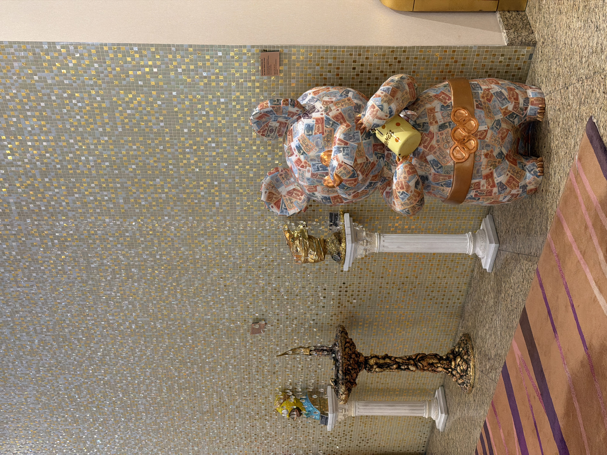 The image shows the interior of a building in Moscow, Russia, featuring art installations. A mosaic wall serves as the backdrop. An art installation of a large teddy bear with a belt and a yellow object in its hand stands on a white pedestal. Below it is another installation of a bronze-like object. A striped rug is visible on the right side of the image.