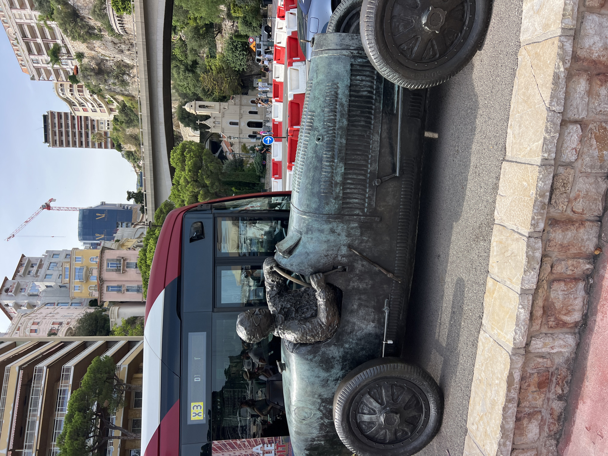An eye-level, medium shot captures a bronze sculpture of a race car and driver in Monaco. The sculpture is positioned on the left side of the frame, facing right, and is predominantly green from weathering. The driver, wearing goggles, has his hands on the steering wheel. A red and white bus is visible behind the sculpture, its side displaying "X3" and other illuminated text. In the background, several buildings with balconies and pastel-colored facades rise up a steep hill. A bridge crosses over the scene, and a small church with a bell tower is visible further in the distance. Red and white traffic barriers are placed in front of the church. The ground is paved with asphalt, and a low stone wall with a red-painted edge runs along the bottom of the frame. The sky is clear and blue.