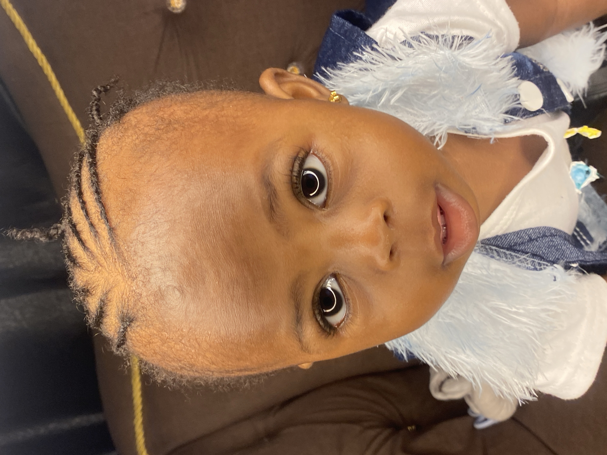 The image shows a close-up, head-on shot of a young child with dark skin and braided hair. The child is wearing a white t-shirt with a blue denim vest adorned with a fluffy white trim around the collar. The child has large, dark eyes and is wearing a small gold earring in their right ear. The background is dark and out of focus, suggesting an indoor setting. The scene is likely within a residential environment in Abakaliki, Nigeria, given the context provided.