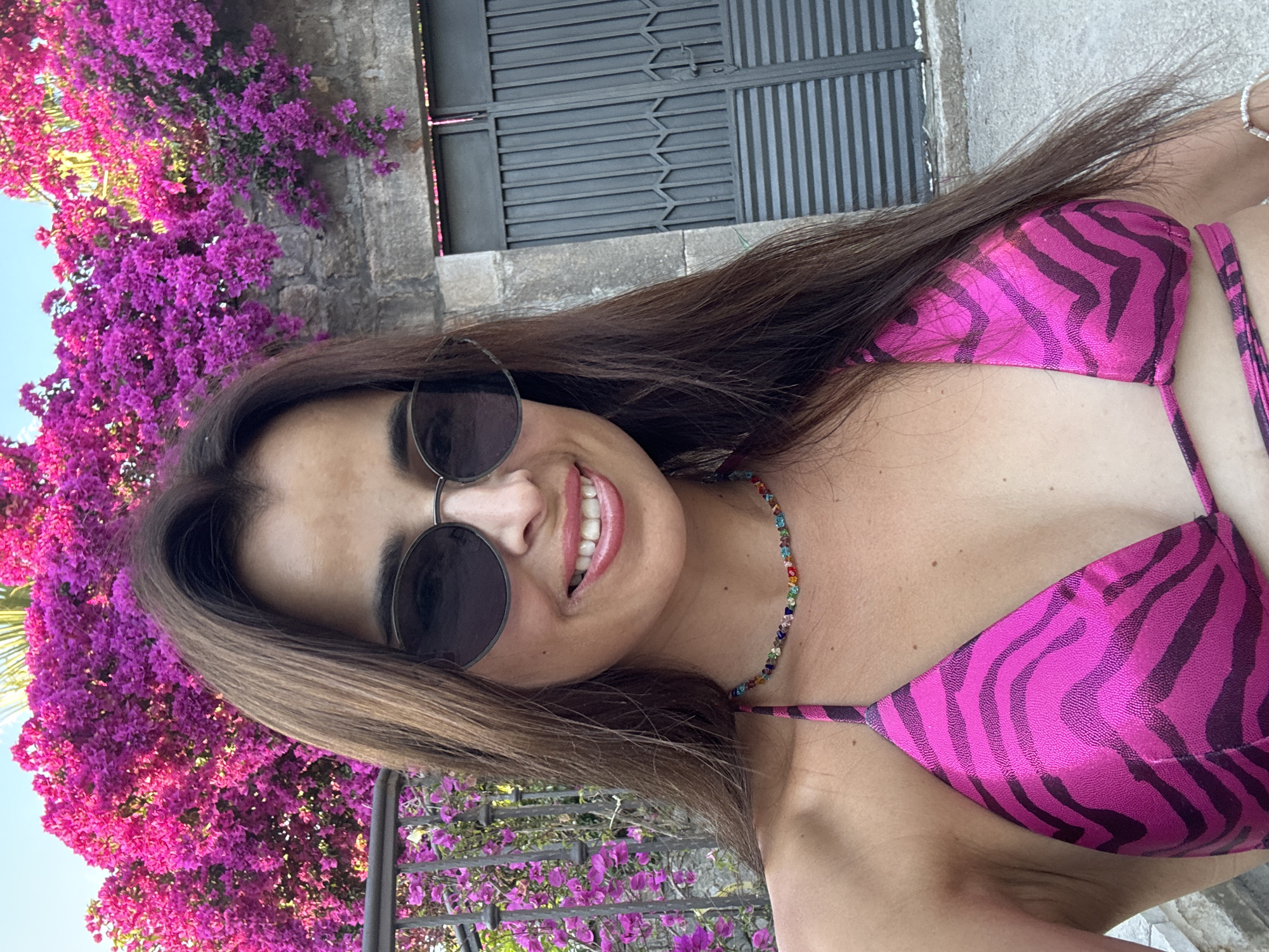 A young woman is captured in a selfie outdoors, with a vibrant cascade of pink bougainvillea flowers to her left. She's wearing a pink and black zebra-striped bikini and a multi-colored beaded necklace. Her long, dark hair is casually draped around her shoulders. She's wearing dark, round sunglasses that partially obscure her eyes, but her smiling face is clearly visible. The background features a stone wall and a dark metal gate. The lighting suggests it is daytime, possibly late morning or early afternoon, with bright sunlight illuminating the scene. The overall mood is cheerful and relaxed, as indicated by her smile.