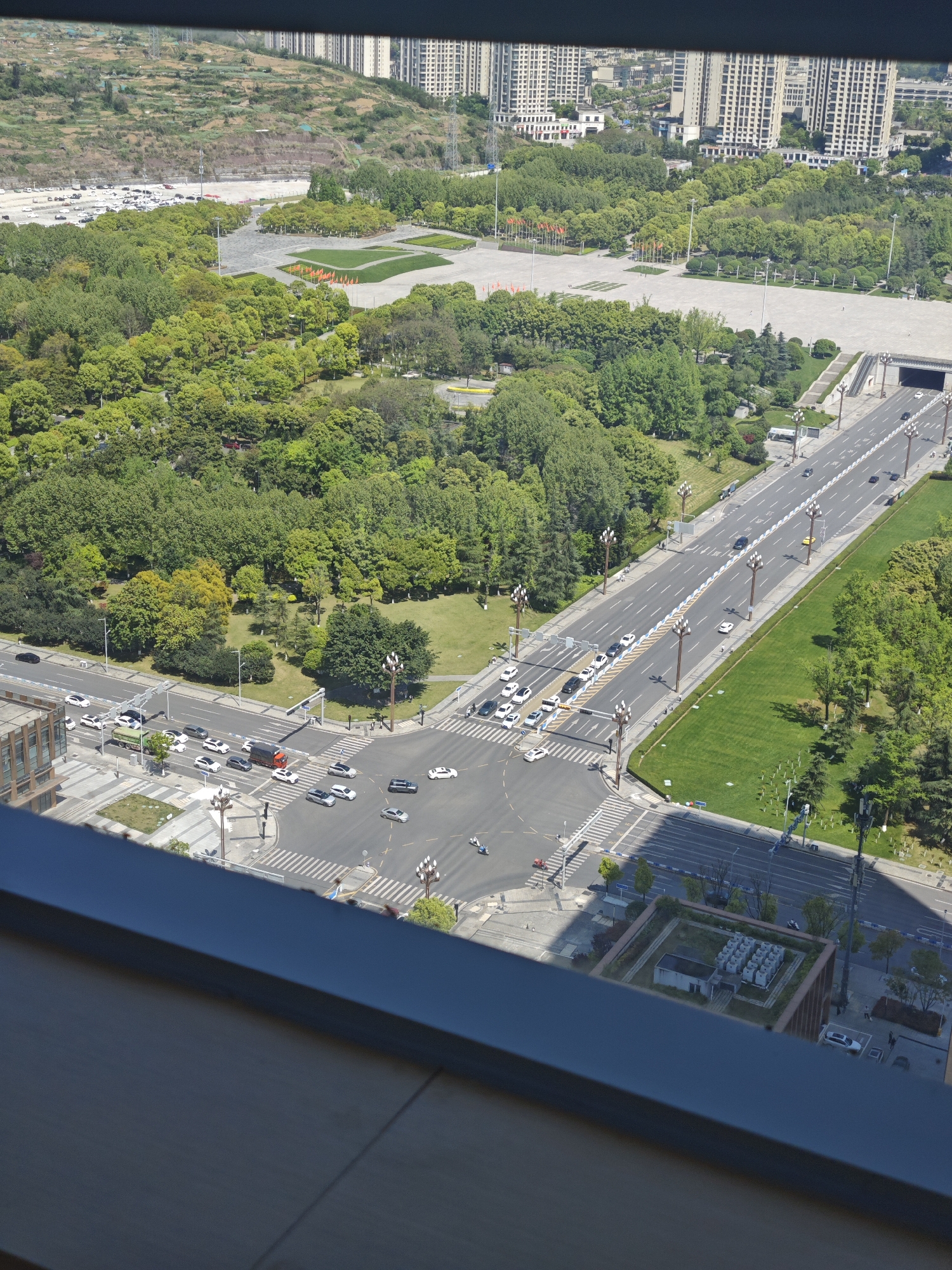An elevated view captures an urban landscape in the Liangjiang New Area of Chongqing, China. A large, multi-lane road bisects the scene, populated by cars and framed by ornate streetlights and manicured green spaces. An intersection with multiple crosswalks is bustling with vehicular and pedestrian traffic. Adjacent to the road is a dense expanse of trees interspersed with grassy areas. Beyond the trees, a vast concrete plaza is visible, adorned with a symmetrical arrangement of flags and landscaping. Further in the distance, a hillside is dotted with low-rise structures and parking lots, leading to a skyline punctuated by high-rise residential buildings. The image is taken through a window, as indicated by the frame along the bottom and left edges. The lighting suggests a clear, sunny day.