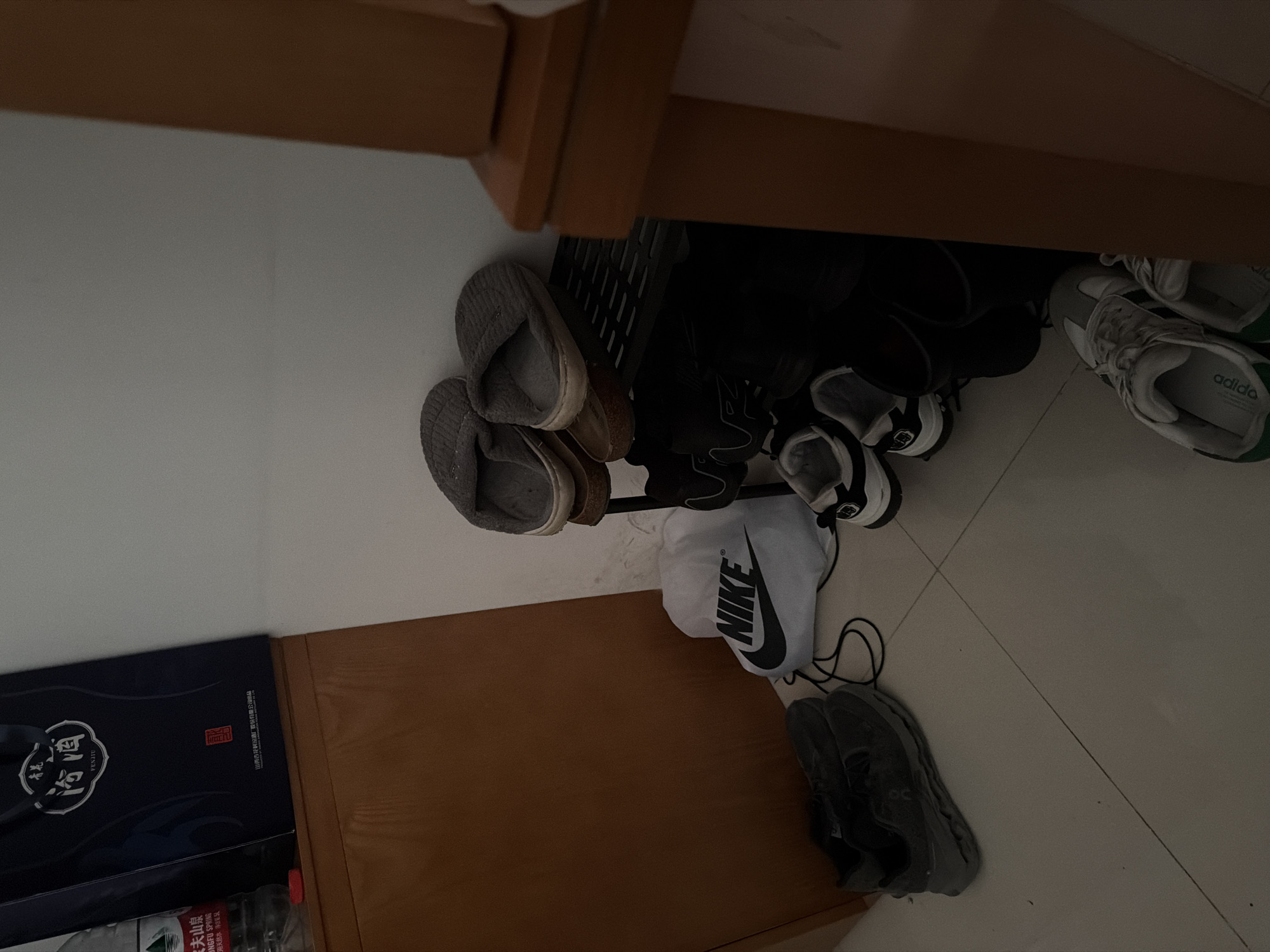This image captures a cluttered storage area, possibly under a piece of furniture like a bed or table. A variety of footwear is visible, including gray slippers, several pairs of black shoes, a pair of gray Adidas sneakers, and gray running shoes. A white Nike drawstring bag is among the shoes. A box of Baijiu, specifically "Fenjiu" is partially visible next to the furniture. The bottles of Nongfu Spring are under the box. Based on the brand names and text in the image, it's plausible that this scene is located in Jiangning District, China.