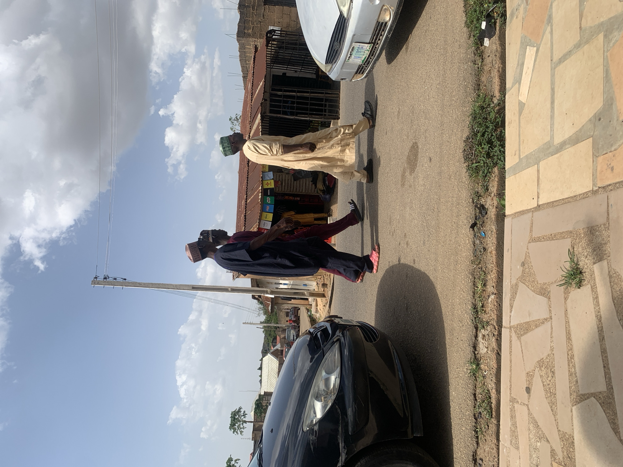 Two men, dressed in traditional Nigerian attire, are crossing a street in Jos, Nigeria. One man wears a light-colored robe and a green cap, while the other wears a maroon robe and a peach cap. A black car is parked on the left side of the street, and a silver car is parked on the right. A small storefront is visible in the background. The street is paved, and there is a sidewalk on the right side. A utility pole with wires is on the left. The sky is blue with scattered clouds.