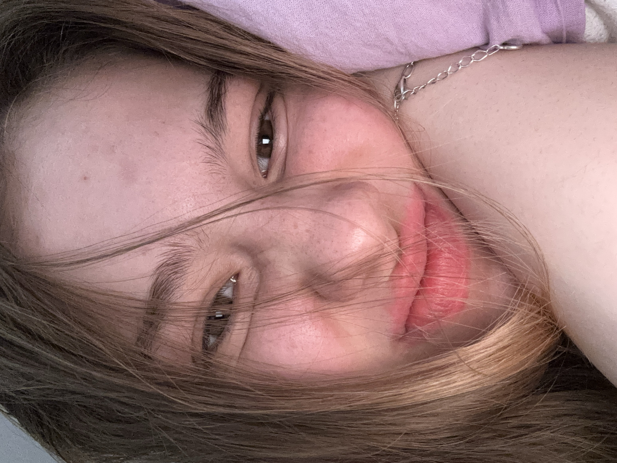 This is a close-up selfie of a young person with light brown hair that is falling across their face. They have brown eyes, light skin with some freckles, and rosy cheeks. They are wearing pink lipstick and a silver chain bracelet. The background is a soft purple color, likely a pillow or blanket, and appears to be indoors. The overall lighting is soft and warm, suggesting it might be natural light or a warm artificial light source. There is no information in the image to suggest a location in Vsevolozhsk, Russia.