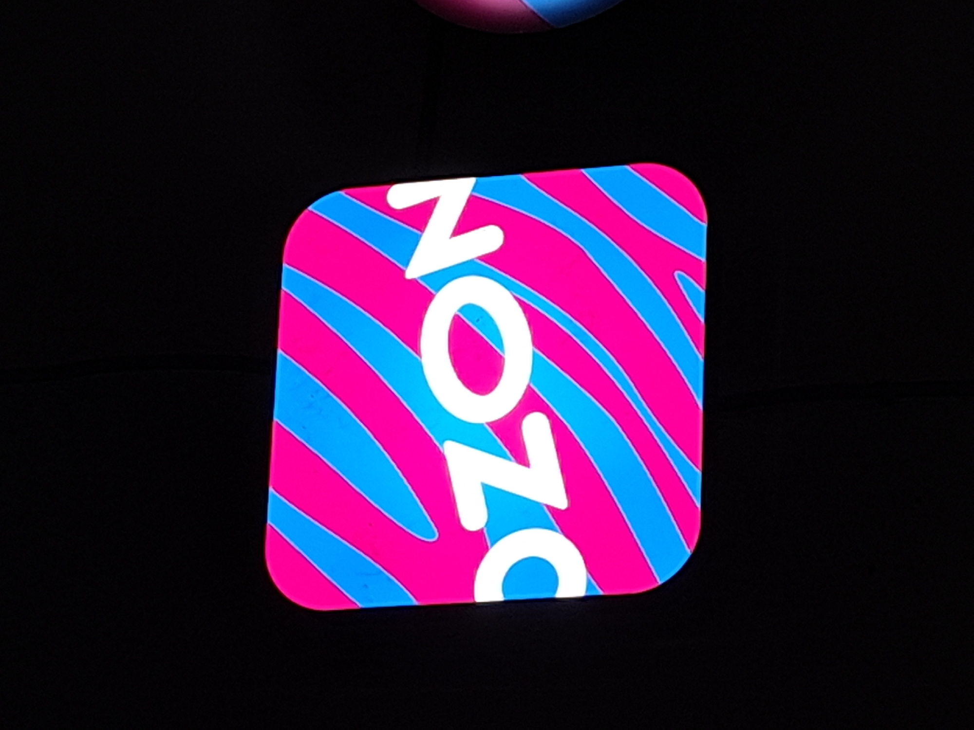 The image displays a close-up of a sign with the word "OZON" in large white letters against a background of pink and blue diagonal stripes. The sign is rounded at the corners and appears to be illuminated. To the right of the sign, a portion of a pink and blue striped sphere-like object is visible. The overall background of the image is black. There is no discernible information in the image to indicate a specific location within the city of Irkutsk, Russia.
