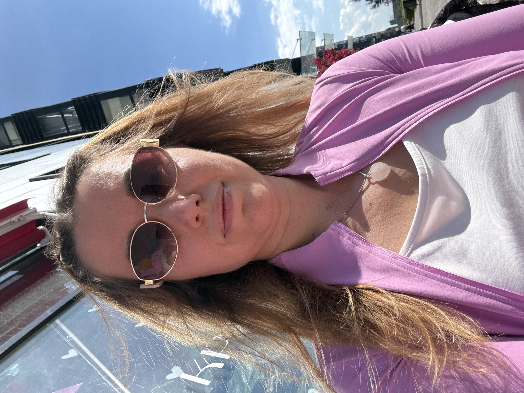 A selfie-style shot captures a woman outdoors on a bright, sunny day. The woman is positioned at an angle, looking up towards the camera. She has long, light brown hair and is wearing large, round, gold-rimmed sunglasses that obscure her eyes. Her lips are slightly parted, revealing a neutral to softly smiling expression.
She is dressed in a light purple or lavender long-sleeved top or jacket, worn open over a white t-shirt. A delicate silver necklace with a light pink, teardrop-shaped stone pendant rests on her chest, visible against the white top.
The background shows a clear blue sky with some wispy white clouds. Modern buildings with dark, reflective glass facades are visible in the distance, suggesting an urban or semi-urban environment. Some vibrant red foliage or flowers add a splash of color to the background. The foreground features a reflective surface, possibly a glass table or barrier, showing indistinct patterns that could be reflections or decorations. The strong sunlight casts clear shadows, indicating it is likely midday or early afternoon. No legible text is visible in the image.