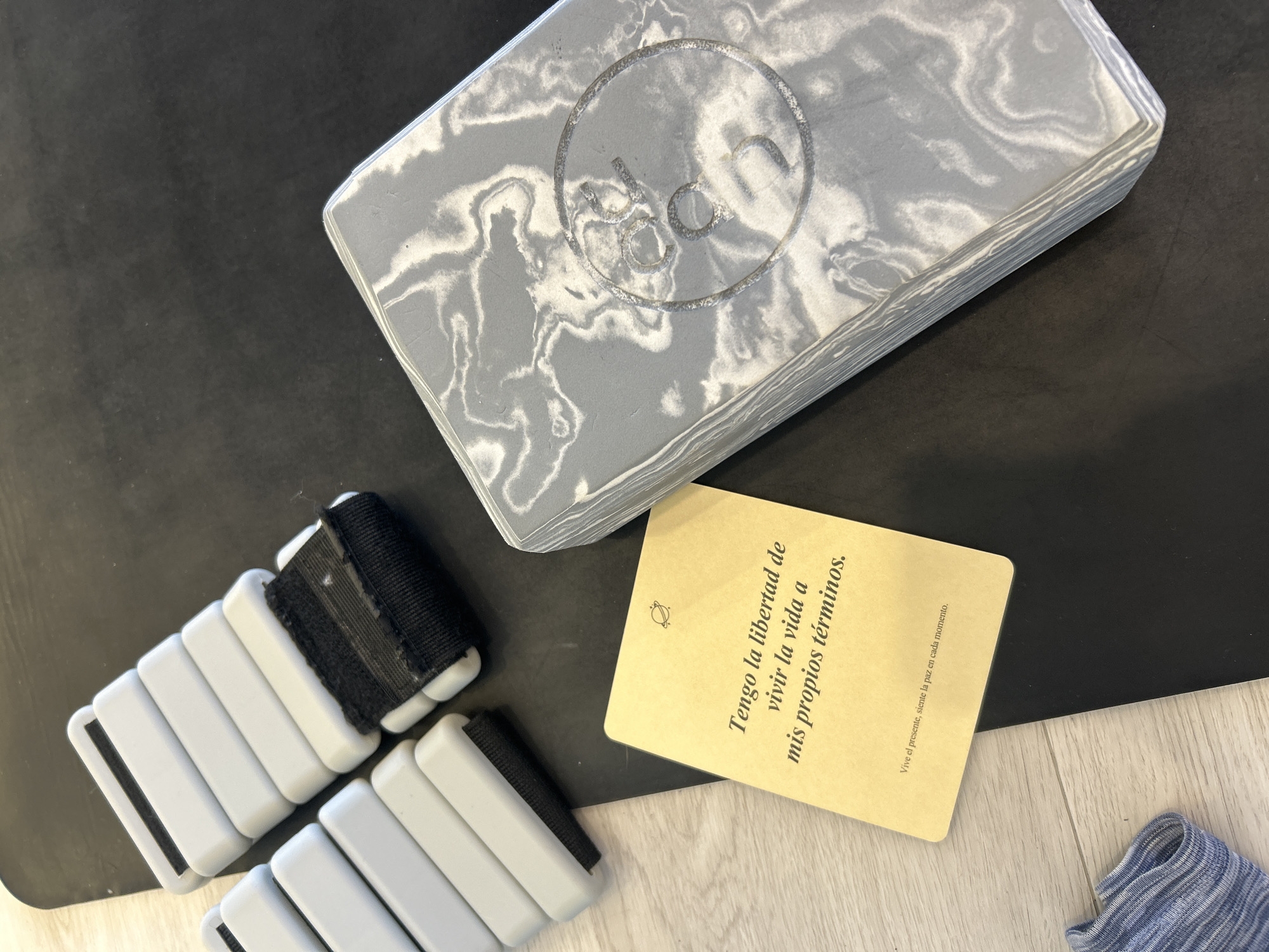 A still life on a dark yoga mat displays items related to yoga or fitness. A gray yoga block with a marbled texture and a circular logo is positioned at the top. To the lower left, two segmented gray ankle weights with black straps are arranged. A beige card with rounded corners, printed with text in Spanish ("Tengo la libertad de vivir la vida a mis propios términos."), sits between the block and the weights. In the lower right corner, a small section of blue and white striped material is visible on a wooden floor.