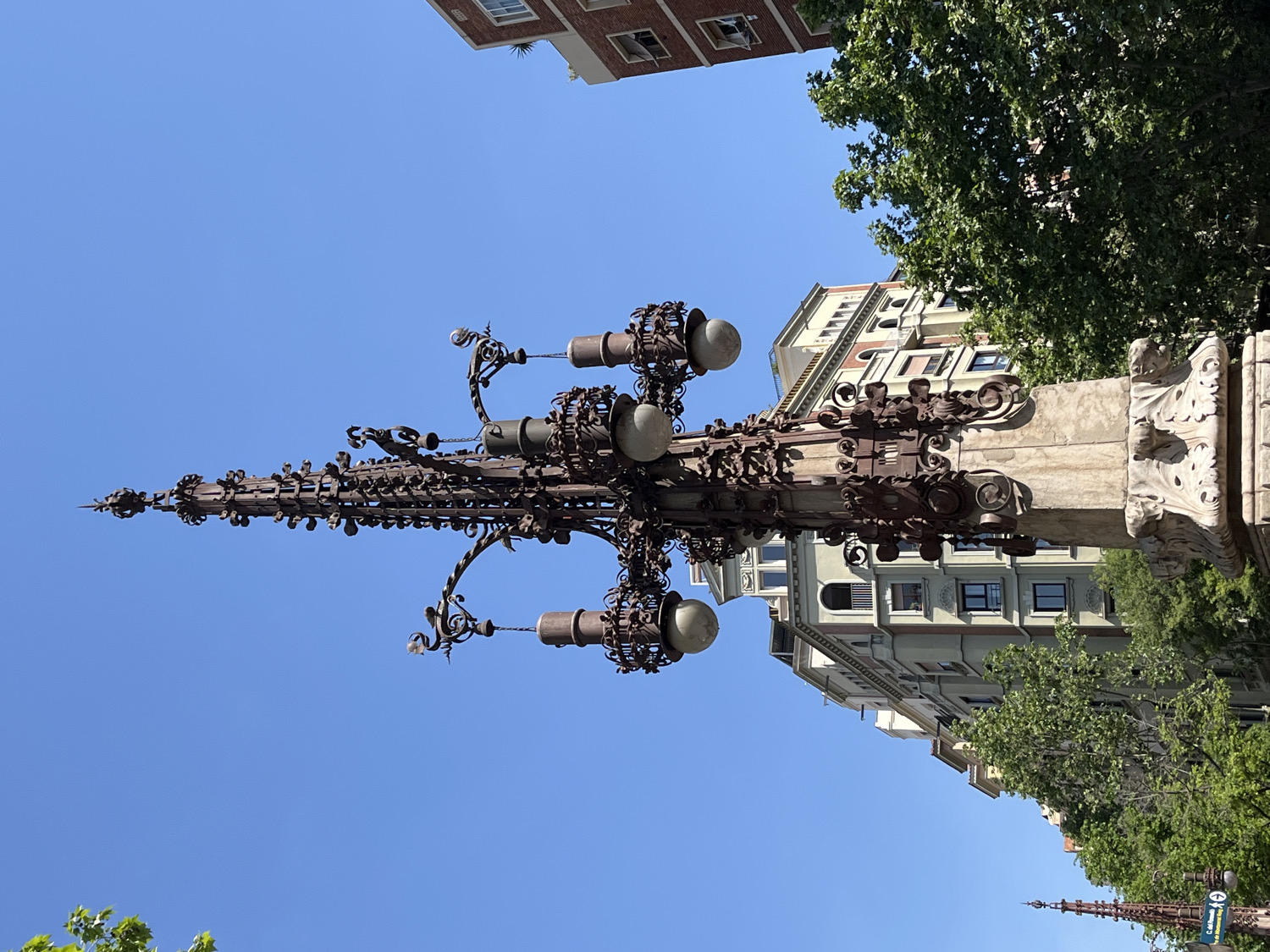 The image displays a highly ornate, dark metal lamppost, designed in a vertical spire-like fashion with intricate ironwork. The lamppost features multiple arms extending outwards, each holding spherical light fixtures. Its upper section is particularly detailed, culminating in a pointed finial, while its base appears to be made of carved stone with decorative flourishes.
The scene is set outdoors under a clear, bright blue sky, indicating sunny weather during the daytime. In the background, several multi-story urban buildings with classic architectural elements like windows and cornices are visible, interspersed with lush green trees. The overall aesthetic of the lamppost and surrounding buildings suggests a European city setting, specifically Barcelona, Spain, given the distinctive modernist-style ironwork reminiscent of the city's architectural heritage.
No specific activity or event is taking place; it's a static street scene. A small street sign is partially visible in the lower right corner, showing the text "Carrer del..." which is Catalan for "Street of the...". The image itself is rotated 90 degrees clockwise from its natural vertical orientation.
