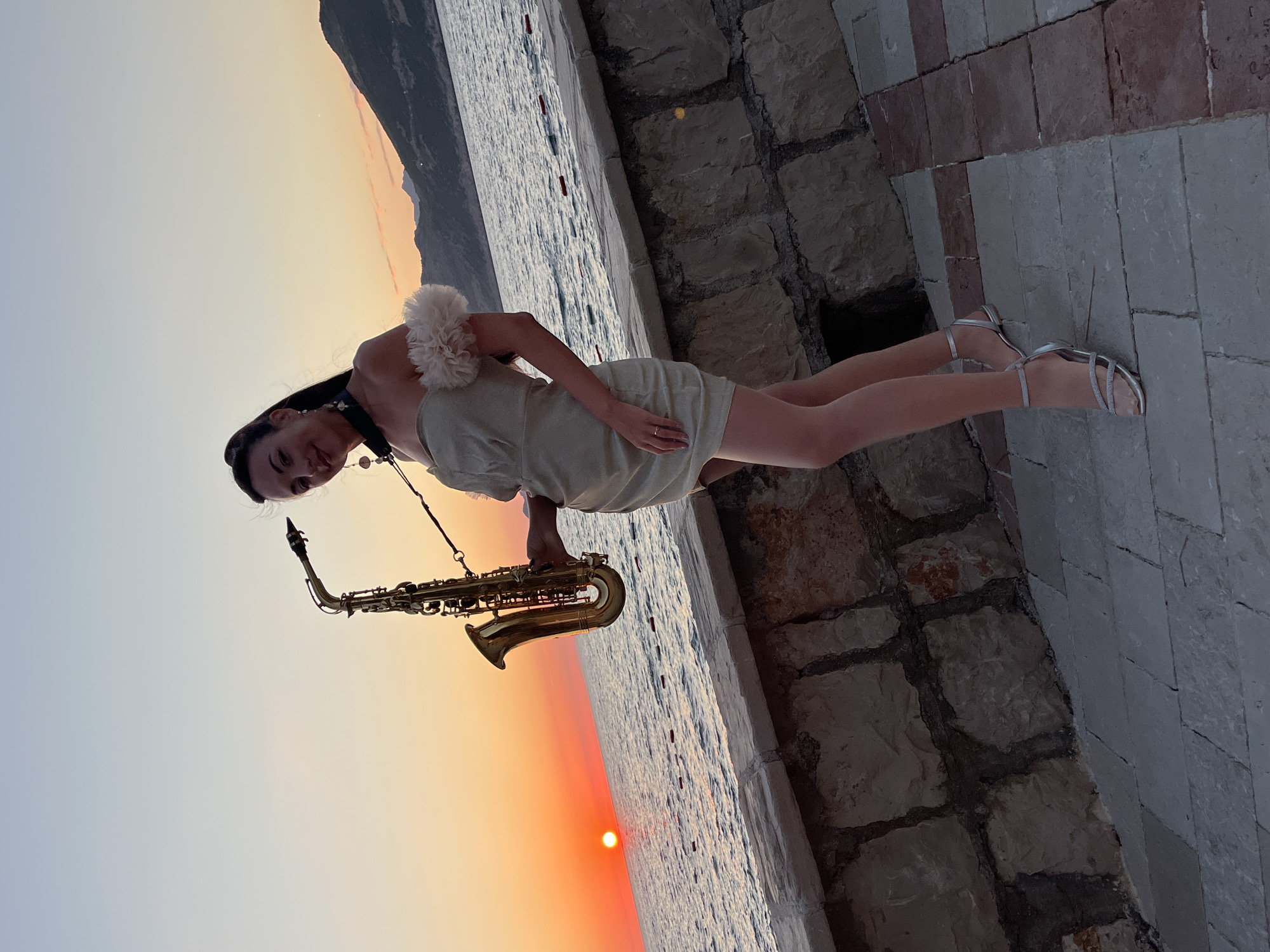 A woman stands on a paved walkway beside a stone wall, holding a saxophone. She is wearing a short, light-colored dress with feathered embellishments on the shoulders and silver strappy heels. The sunset is visible over the sea, with the sun appearing as a bright orange disc low on the horizon. Mountains are visible in the distance. The walkway is paved with stone tiles of varying colors, and the wall is constructed from rough-hewn stones. The scene is outdoors, likely in a coastal area. The approximate location is Pečurice, Montenegro.