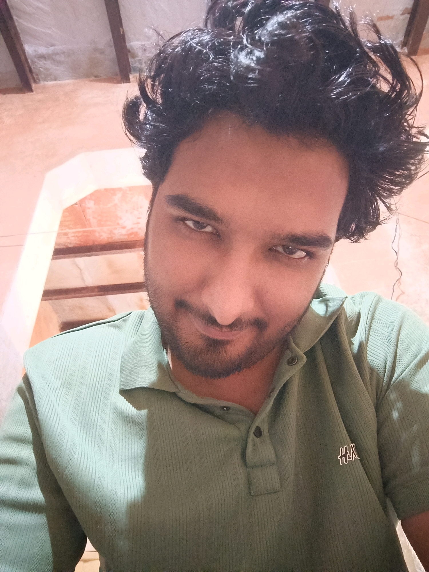 A close-up, waist-up, selfie-style photograph shows a young man with dark, curly hair and a beard. He is wearing a light green, collared, short-sleeved polo shirt with a ribbed texture and two dark buttons at the neck. A small, white logo with black outline, appearing to be "H&M", is visible on the left side of his chest. The man is looking directly at the camera with a slight smile and a direct gaze. The background is out of focus but appears to be an interior space with an orange-toned wall and architectural elements that suggest a building with a staircase or tiered structure. The lighting creates a shadow on the man's right side. The overall impression is casual and personal.