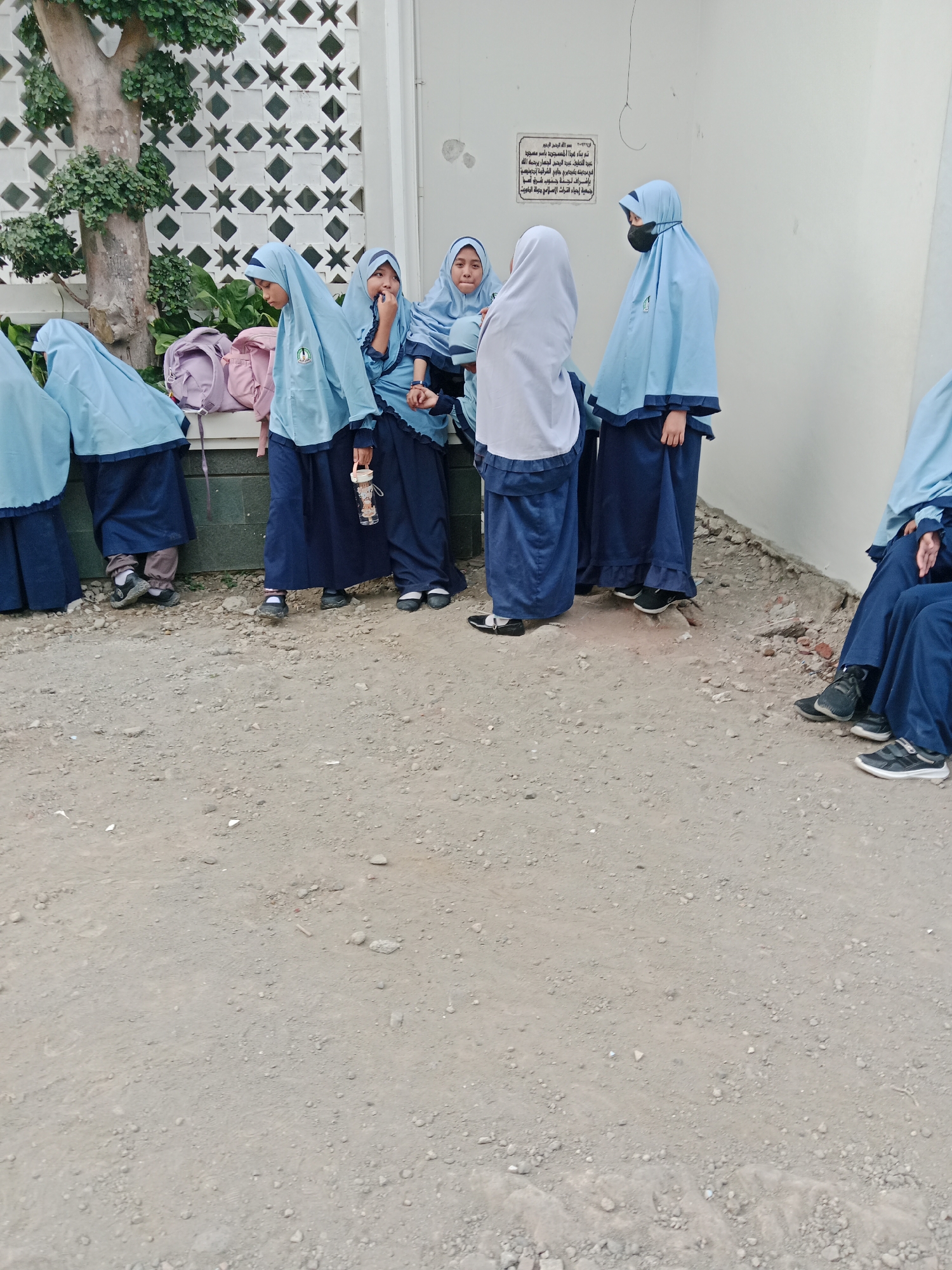 In the city of Kediri, Indonesia, a group of young girls wearing light blue hijabs and dark blue uniforms are standing near a white wall with an ornate patterned partition. Some girls have their backpacks with them, one pink and another lilac. The ground is covered in dirt and gravel. One girl is wearing a black mask over her mouth and nose. A small sign with Arabic script is affixed to the white wall.
