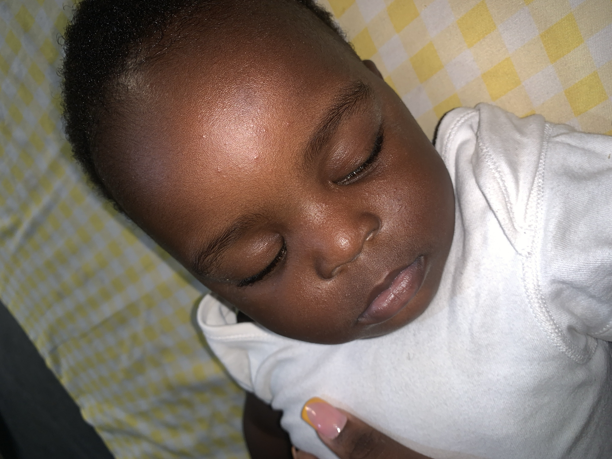 A close-up, overhead shot shows a sleeping Black baby wearing a white onesie. The baby's face is turned to the right, with eyes closed and lips slightly parted. A portion of a person's hand with a light pink and yellow manicured nail is visible on the left side of the image, touching the baby's onesie. The baby is lying on a yellow and white checkered surface. The lighting is dim, casting a warm glow on the baby's skin.