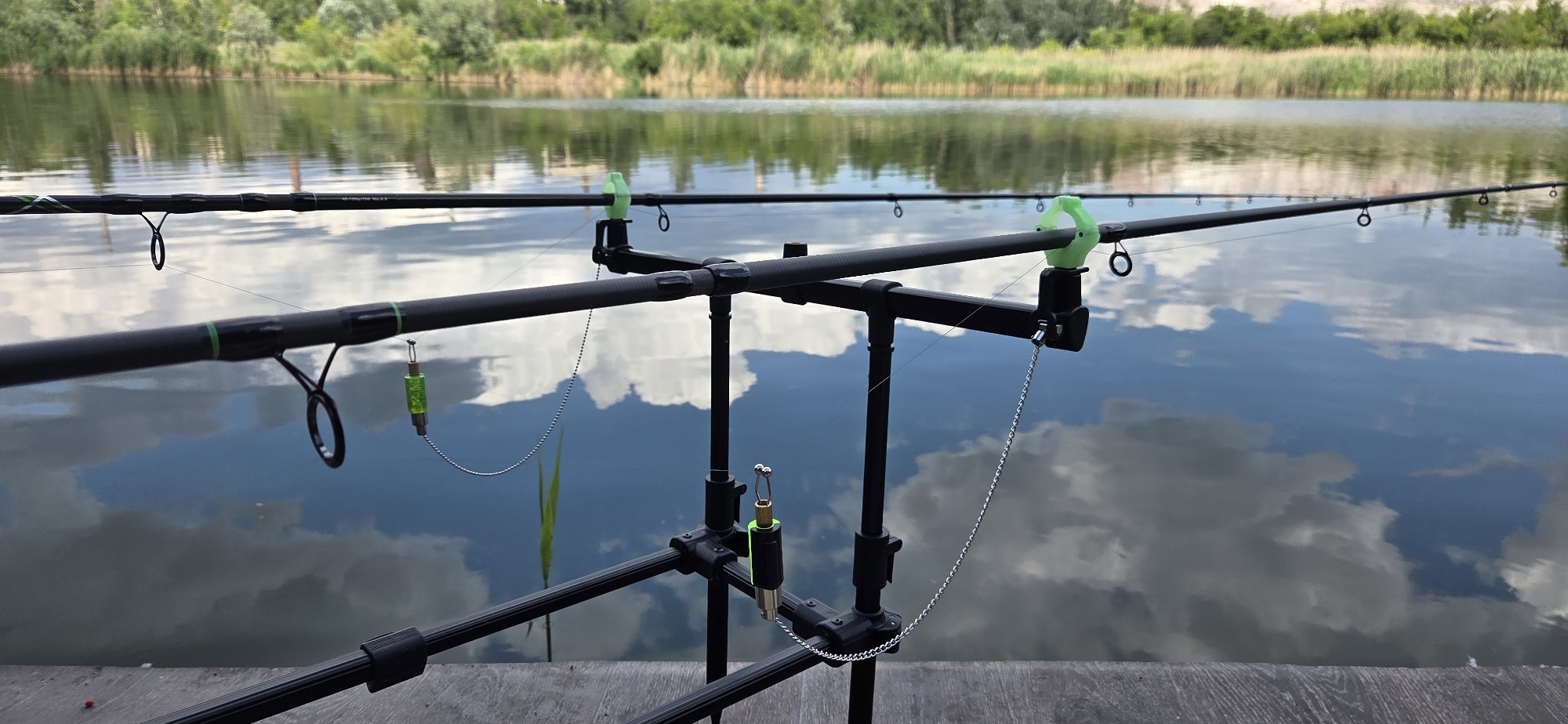 The image shows a close-up view of two fishing rods set up on a rod pod beside a calm lake. The fishing rods are black with green accents and have multiple guides. The rod pod is also black and appears to be made of metal. On each rod, a fishing line extends out towards the water, and a green fishing bite indicator is attached. One bite indicator is cylindrical and translucent green, while the other is shorter and wider, also with a green top. Both are suspended by thin metal chains.
The lake's surface reflects the sky, which is partly cloudy, suggesting a bright but not entirely sunny day. The water is still, creating a mirror-like effect. In the background, there is a dense line of reeds and trees along the far bank of the lake, with a lighter colored area that might be a hill or buildings beyond. The foreground shows the edge of a wooden deck or pier. There is no visible text in the image. The scene appears to be set up for fishing, with the rods ready to be used, implying an activity of fishing is either about to commence or is in progress. The overall mood is tranquil and peaceful, characteristic of a day spent by the water.