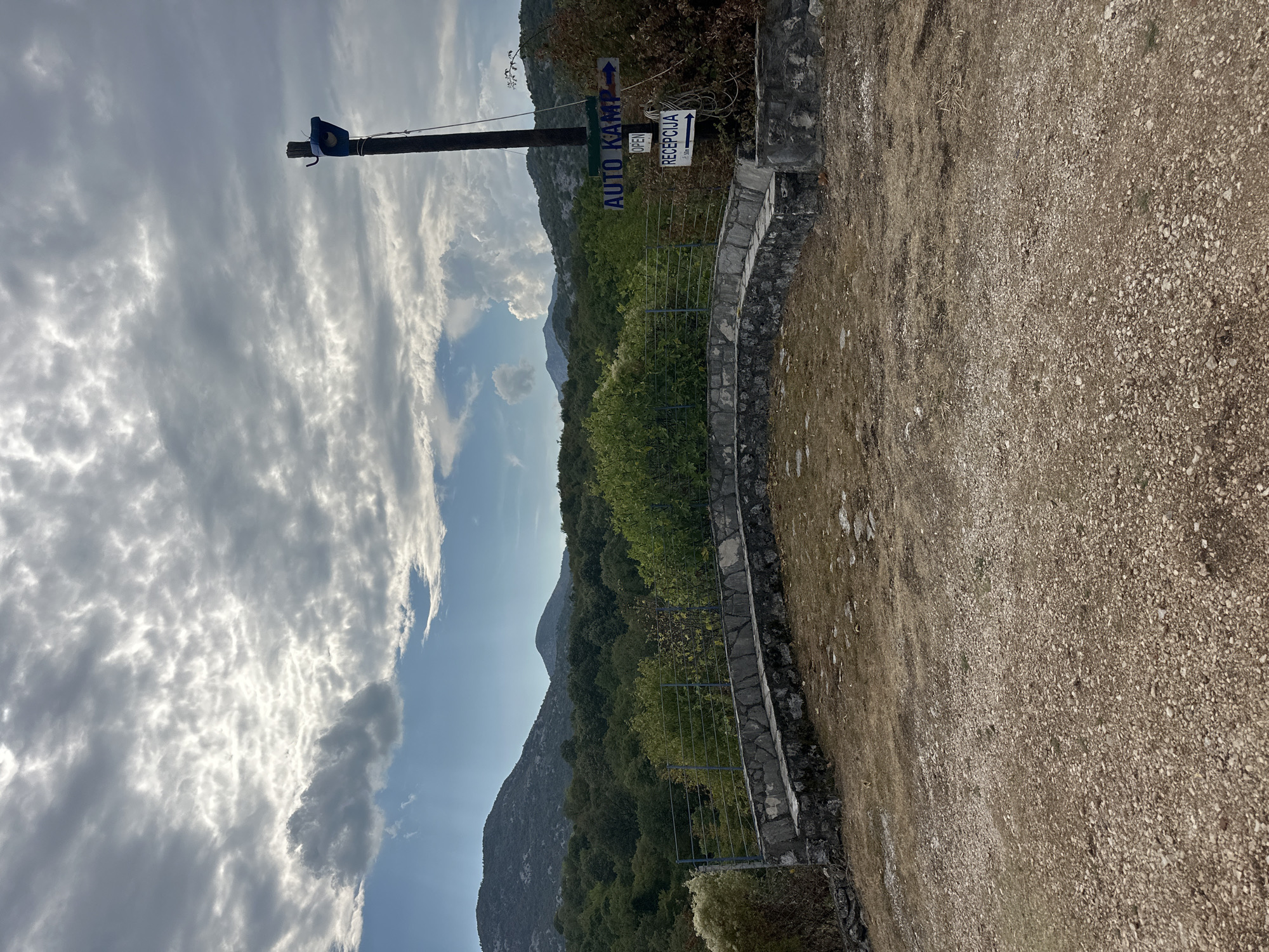 The image depicts an outdoor scene, likely a campsite, in Cetinje, Montenegro. In the foreground, a gravelly area leads to a low stone wall and a tiered seating area. Behind this, a metal fence encloses a patch of dry grass and vegetation.
A prominent wooden pole stands to the right, topped with a blue object and a string. Attached to the pole are two blue and white directional signs. The upper sign reads "AUTO KAMP" with an arrow pointing to the right, and below it, the word "OPEN" is visible. The lower sign says "RECEPCIJA" with an arrow also pointing to the right and a distance marker of "50m".
The background features a dense forest of green trees, with rolling hills and mountains further beyond under a cloudy sky. The lighting suggests it might be late afternoon or an overcast day. No people are visible in the frame.