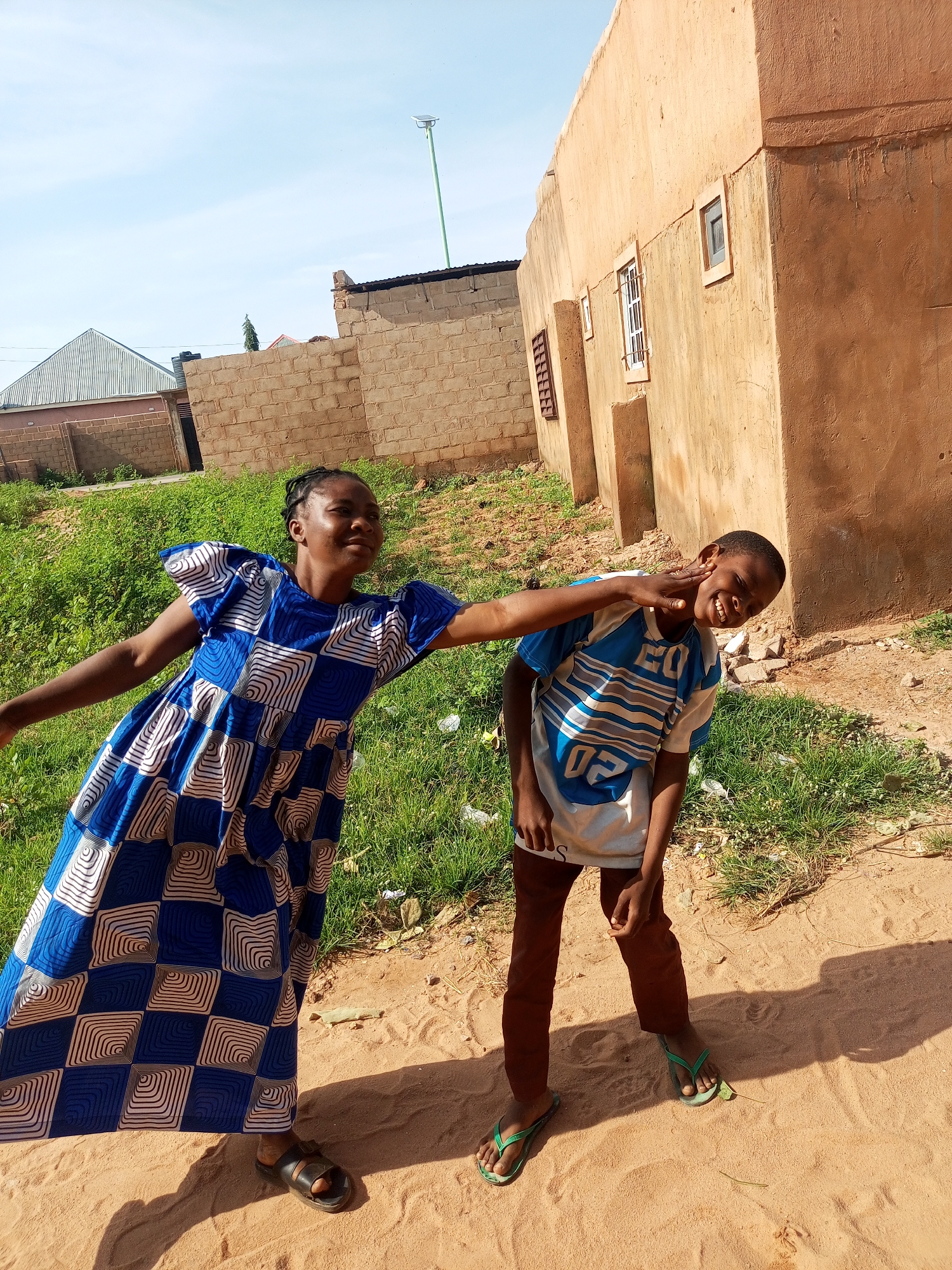 Two children are standing in an outdoor, sandy area with sparse green grass and a dirt path. In the background, there are buildings constructed from light-colored bricks and a larger, rust-colored building with windows. A young girl on the left is wearing a blue and white patterned dress and black sandals. She has her right arm extended and her hand resting on the face of a younger boy to her right. The boy is wearing a white t-shirt with blue stripes and the numbers "02", dark brown pants, and green flip-flops. He is smiling and tilting his head back as if being tickled or playfully pushed. The sky is light blue with some faint clouds.
