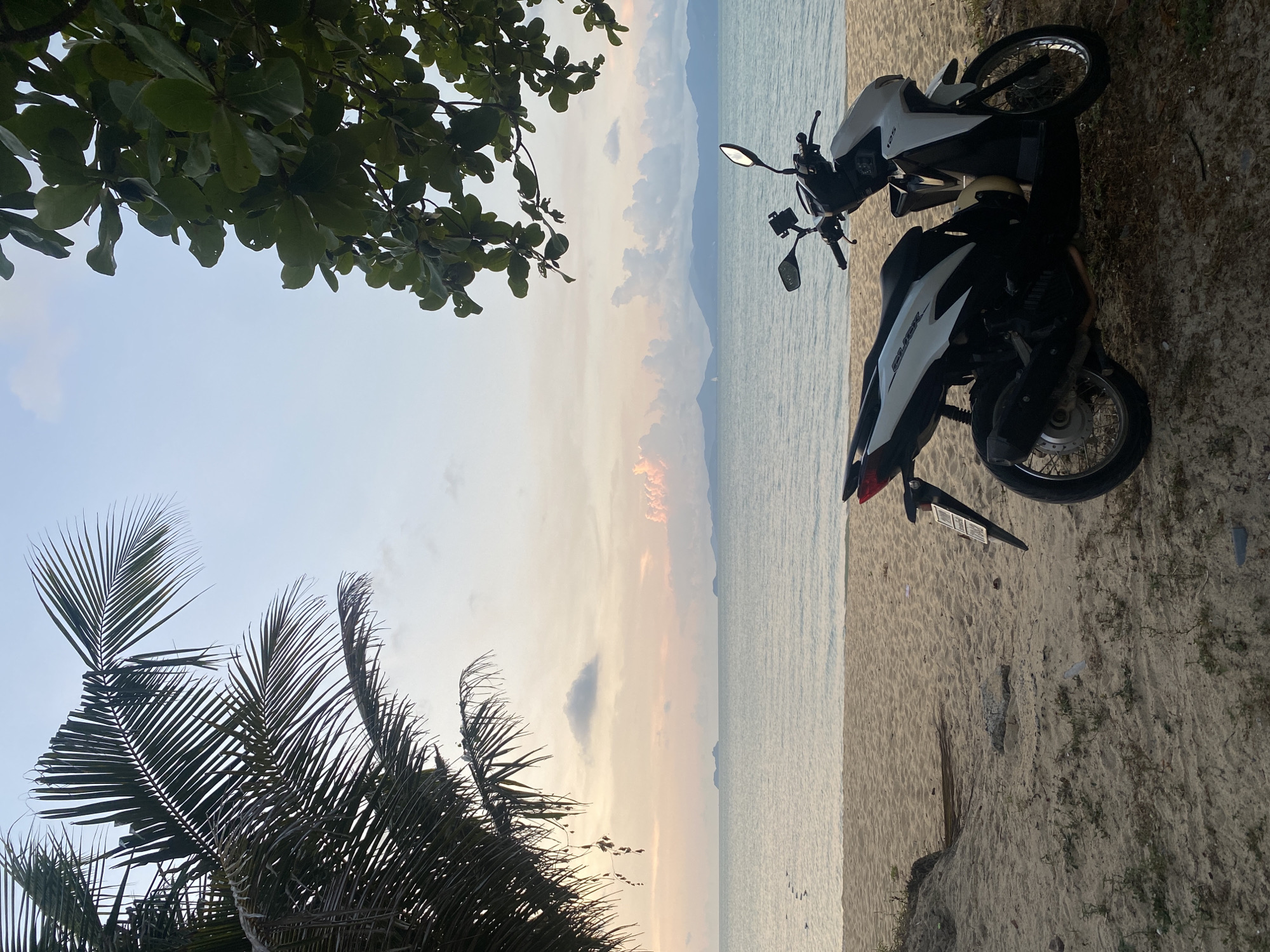 The image depicts a coastal scene in Baan Bo Phut, Thailand at sunset. Two motorcycles are parked on the sandy beach. The sea stretches to the horizon, with silhouetted islands visible in the distance. The sky is a blend of colors ranging from pale blue to orange. Trees frame the top and left side of the image. The environment is calm and serene, suggesting a quiet beach setting. There are no people visible in the image.