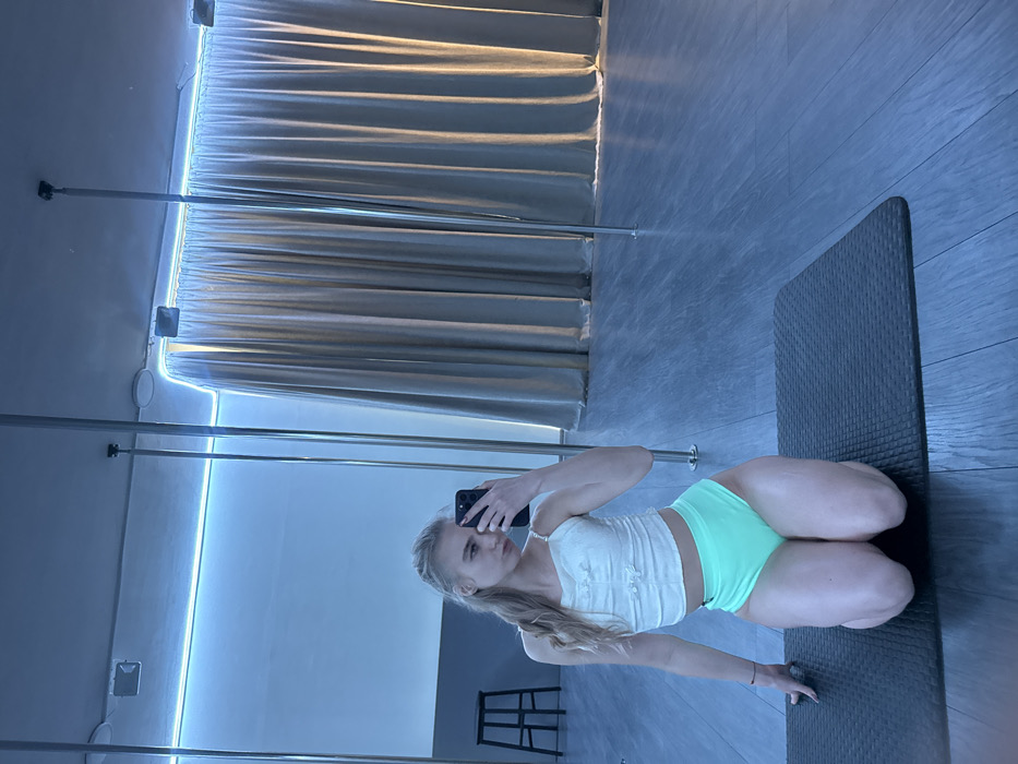 The scene depicts a young female in a pole fitness studio. She is light-skinned with blonde hair, styled in a braid over her left shoulder. She is kneeling on a dark grey exercise mat, wearing a white, ribbed crop top with thin straps and light green, high-waisted briefs. Her right hand holds a black smartphone, taking a mirror selfie, while her left hand rests on the mat. She looks directly towards the phone. The background consists of a mirrored wall reflecting her image and the studio. Visible within the reflection are multiple chrome vertical poles extending from the floor to the ceiling, characteristic of pole dance equipment. A blue LED strip light illuminates the upper section of a wall. The floor is covered with light grey, wood-look laminate planks. In the far left background, a dark, simple high stool is visible. To the right, light-colored drapes or curtains are present. The specific location within the city of Unknown, Unknown, cannot be determined from the image.