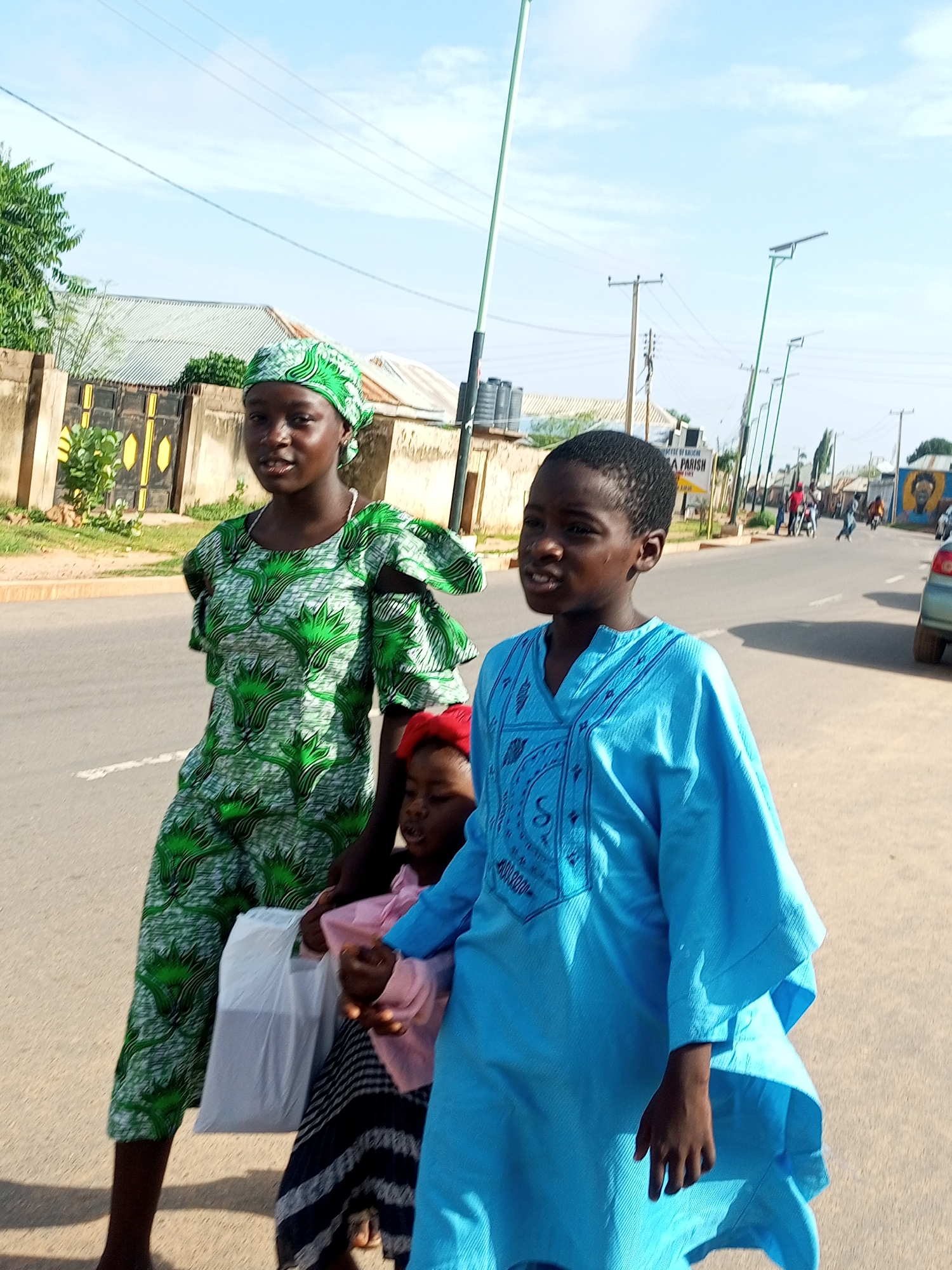 In Gombe, Nigeria, on a paved road under a clear sky, three individuals are captured in a medium shot. A young woman in the foreground, on the left, wears a green and white patterned dress and headscarf. To her right, a child in a pink top and a striped skirt is held by another individual, partially visible, wearing a bright blue tunic. The background reveals a street with parked cars, pedestrians, and buildings. Power lines and streetlights are visible overhead, indicating an urban setting.