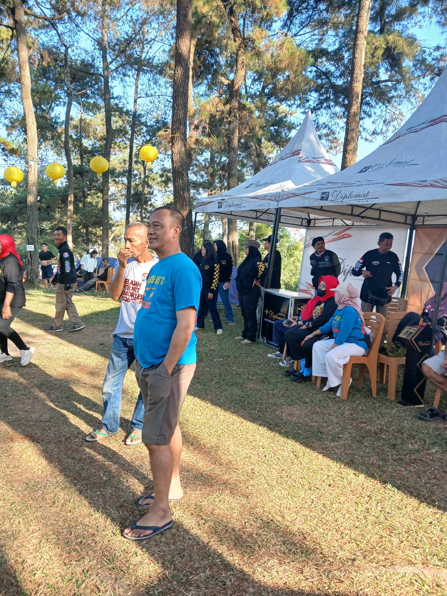 The image depicts an outdoor gathering in the city of Berjo, Indonesia, likely during daylight. The scene is set in a park-like environment with tall trees, some with autumn-colored foliage, and dry grass covering the ground. The sunlight casts distinct shadows across the lawn, indicating a clear sky.
In the foreground, two men are standing, facing away from the camera and towards a gathering under a tent. The man on the right wears a bright blue t-shirt and khaki shorts, with his hands in his pockets. The man to his left wears a white t-shirt and blue jeans, and appears to be eating or chewing something.
Behind them, a white tent with red accents and the brand name "Diplomat" is set up, suggesting a promotional event or a temporary stall. Several people are gathered under and around this tent, some seated on chairs, others standing. They are dressed in casual clothing, with some individuals wearing headscarves.
In the background, more trees and a blue sky are visible. There are also yellow lanterns hanging from strings between trees, adding a decorative element to the scene. Other individuals are seen walking or standing further away in the grassy area, suggesting a larger event or gathering. The overall atmosphere appears relaxed and social.