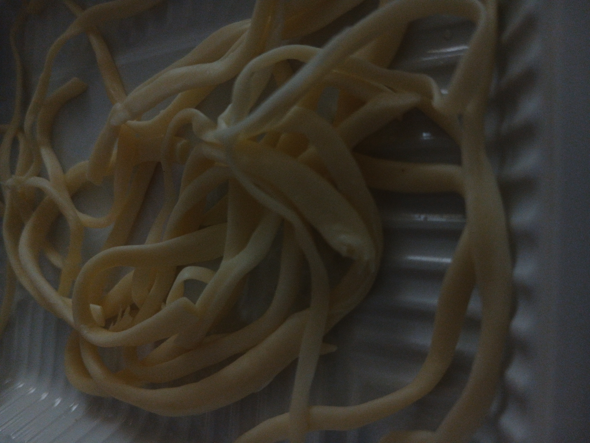 The image shows a close-up, overhead view of a pile of uncooked, thick, yellow noodles arranged in a loose tangle within a white plastic container. The noodles appear to be a type of pasta, possibly bucatini or perciatelli, given their tubular shape and thickness. The container has a ribbed surface on the bottom. The lighting is dim, casting soft shadows and highlighting the textured surface of the noodles. The background is a muted gray, with vertical lines from the container's ridges visible. There are no people or animals in the image, and no discernible activity is taking place. The scene is static and focuses solely on the uncooked pasta. The location in the city of Kasakh, Armenia, is not discernible from the visual information provided in the image.