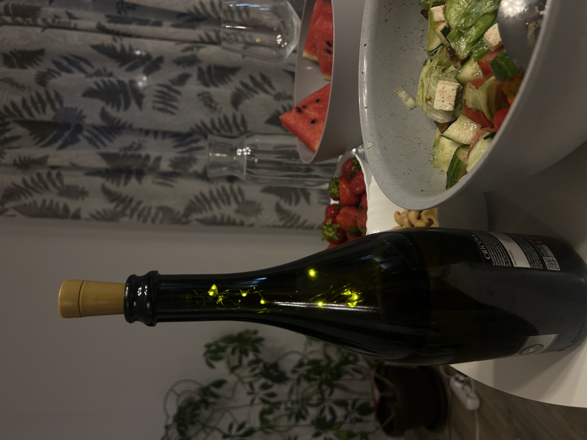 The image depicts a table setting with a bottle of Cricova wine, a bowl of salad, a bowl of strawberries, and slices of watermelon. The bottle of wine is dark green and has small yellow fairy lights inside, creating a festive atmosphere. The salad appears to be a mixed green salad with cucumber, tomatoes, and feta cheese. The strawberries are a vibrant red and look fresh. The watermelon slices are triangular and have visible seeds. In the background, there is a carafe of water and a wine glass, as well as a potted plant and a patterned curtain. The overall scene suggests a casual gathering or a meal enjoyed at home. The location of Florești, Romania, is mentioned as part of the context, but the image itself does not provide any specific visual cues that would confirm or deny this location.