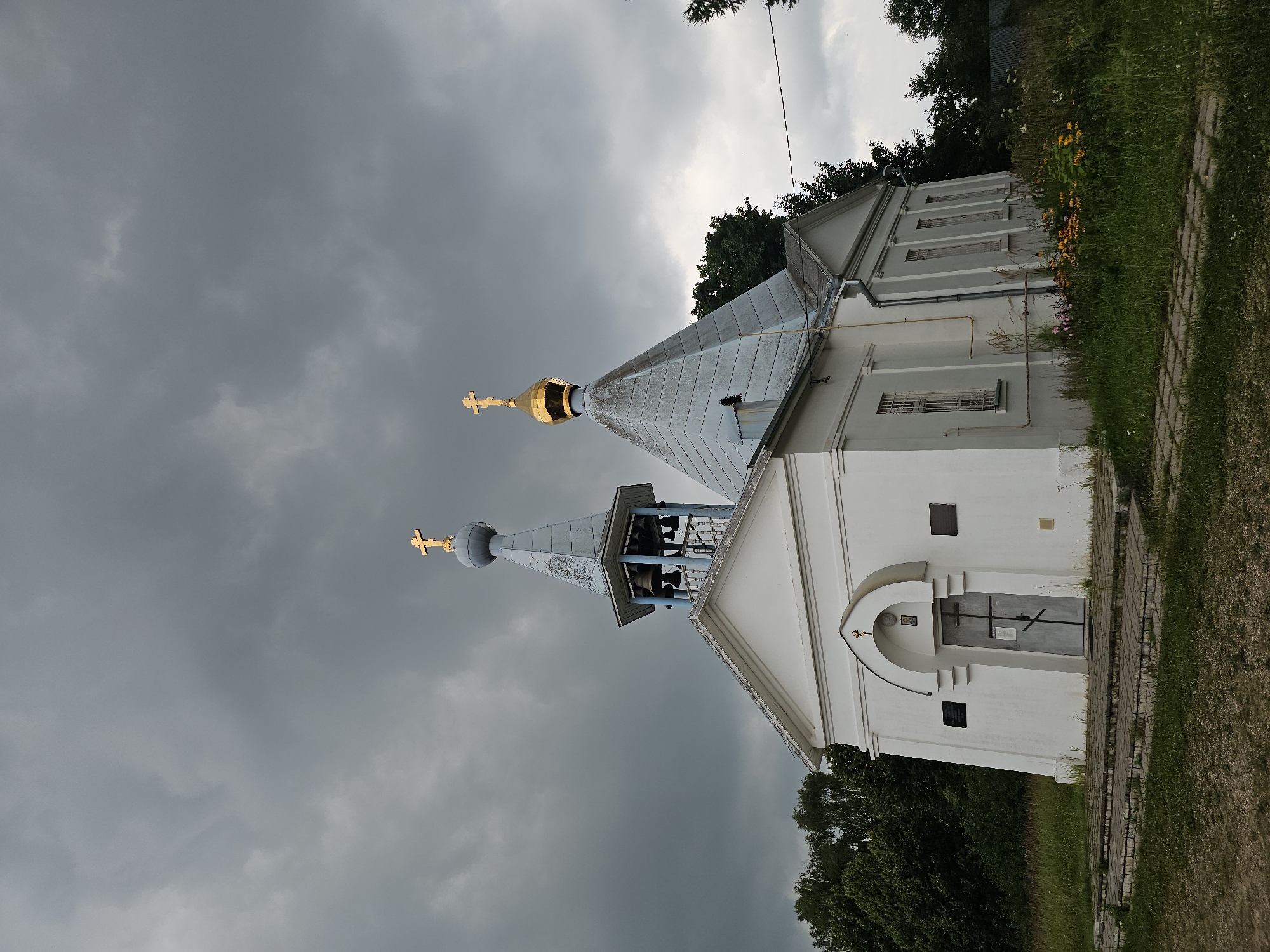 A white, Orthodox church with a bell tower and a gold dome stands against a dark, cloudy sky in Ulitkino, Russia. The church has a grey, pitched roof and a grey door with a cross above it. The bell tower has multiple bells and a golden cross on top. The church is surrounded by green grass, trees, and a few flowers. The ground in front of the church is paved with stone slabs.