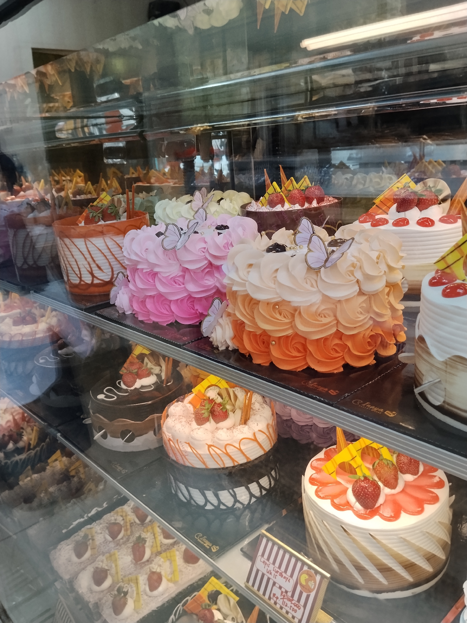 This image captures a display of various cakes inside a glass-fronted bakery cabinet. The cakes are arranged on multiple shelves, with a variety of decorations including frosting rosettes in pink and orange hues, strawberries, and decorative chocolate elements. Some cakes have edible butterfly decorations. In the foreground, a rectangular cake is visible, covered in powdered sugar and topped with sliced strawberries and yellow decorative sticks. A price tag is placed in front of this cake, which appears to be written in Indonesian. The bakery appears to be located in Ciruas, Indonesia, given the presence of Indonesian text on the price tag. The lighting suggests an indoor setting, likely a commercial establishment.