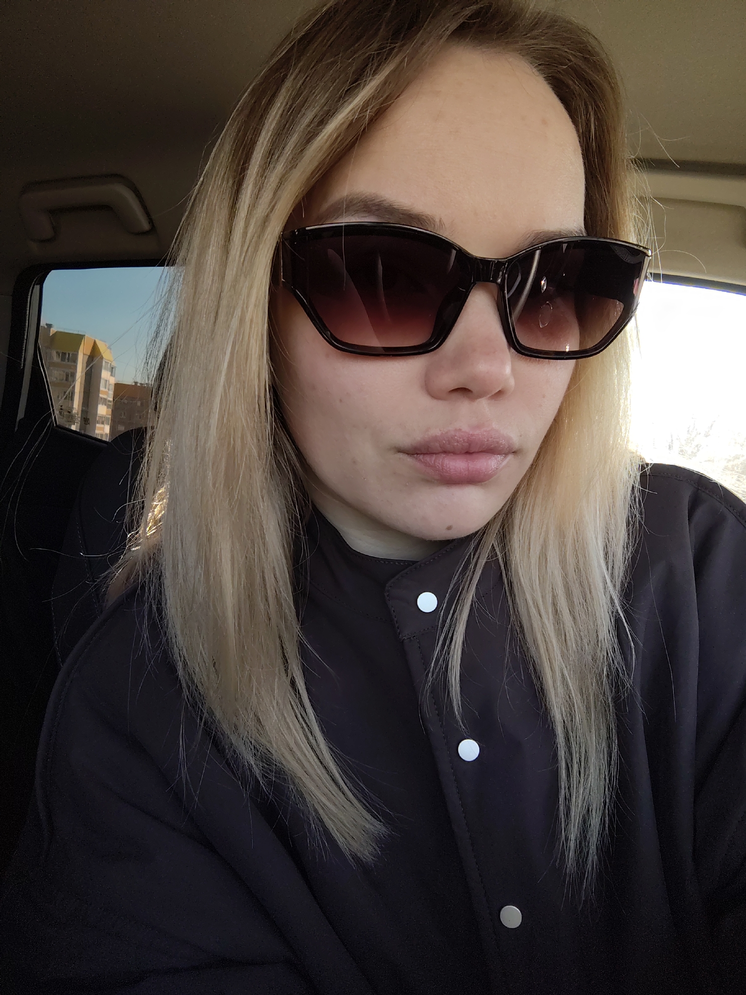 A close-up portrait shows a woman with long, straight blonde hair, wearing dark, stylish sunglasses and a dark brown or black jacket with light-colored snap buttons. She is seated inside a car, likely taking a selfie, with parts of the car's interior visible, including the headliner and a grab handle. Her expression is neutral, partially obscured by the sunglasses, which reflect some light.
Through the car window, an urban or suburban landscape is visible, featuring multi-story residential buildings with some yellow accents on their facades, under a clear, bright blue sky. The strong sunlight casts highlights on her hair and illuminates the scene outside, suggesting it is daytime, possibly morning or late afternoon. The overall impression is a casual moment captured in Markovskiy, Russia. No visible text is present in the image.